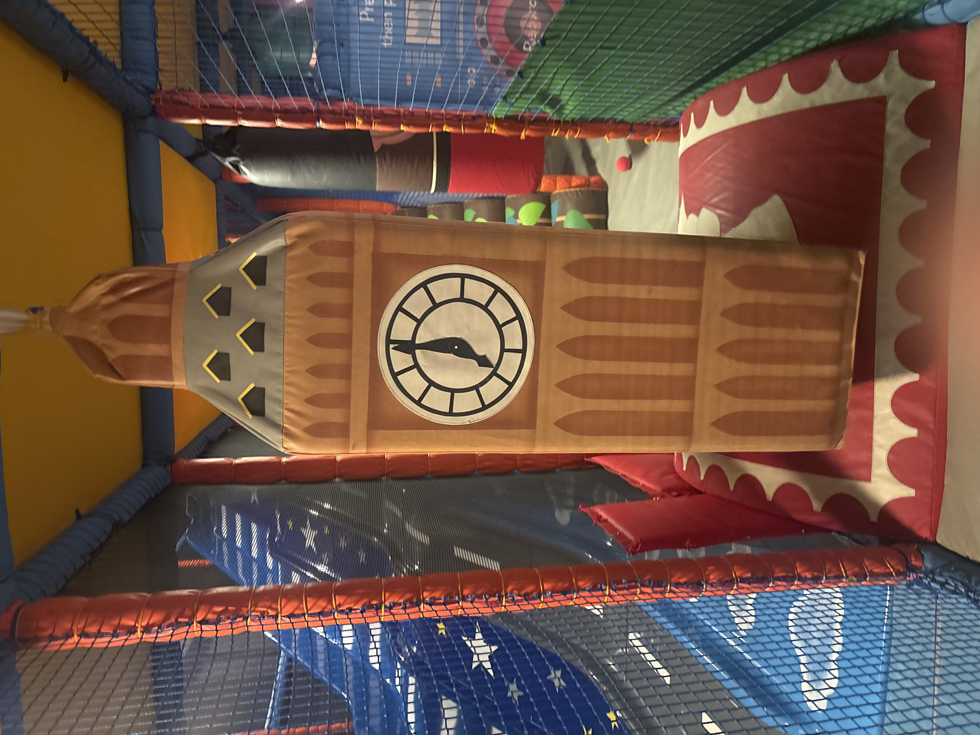 An indoor soft play area features a large, inflatable structure resembling Big Ben. The Big Ben replica has a clock face, with the hands indicating approximately 6:55. It is positioned on a red and white padded mat. Surrounding the central attraction are various play structures, including a blue slide with white star patterns and netting enclosures with orange padding. In the background, a black and red cylindrical inflatable is visible, along with glimpses of green netting and other soft play elements. A small red ball lies on the floor near the Big Ben structure. The ceiling of the play area is a bright yellow fabric. The environment is brightly lit, typical of an indoor children's entertainment facility. No people are visible in the image.