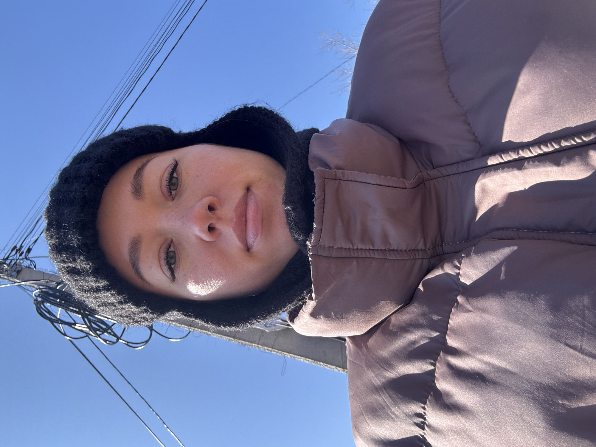 A woman is looking downward at the camera while standing outdoors on a sunny day in Ферма, Russia. She is wearing a black knit hood and a brown puffy jacket. In the background, there is a clear blue sky and some utility poles with wires.