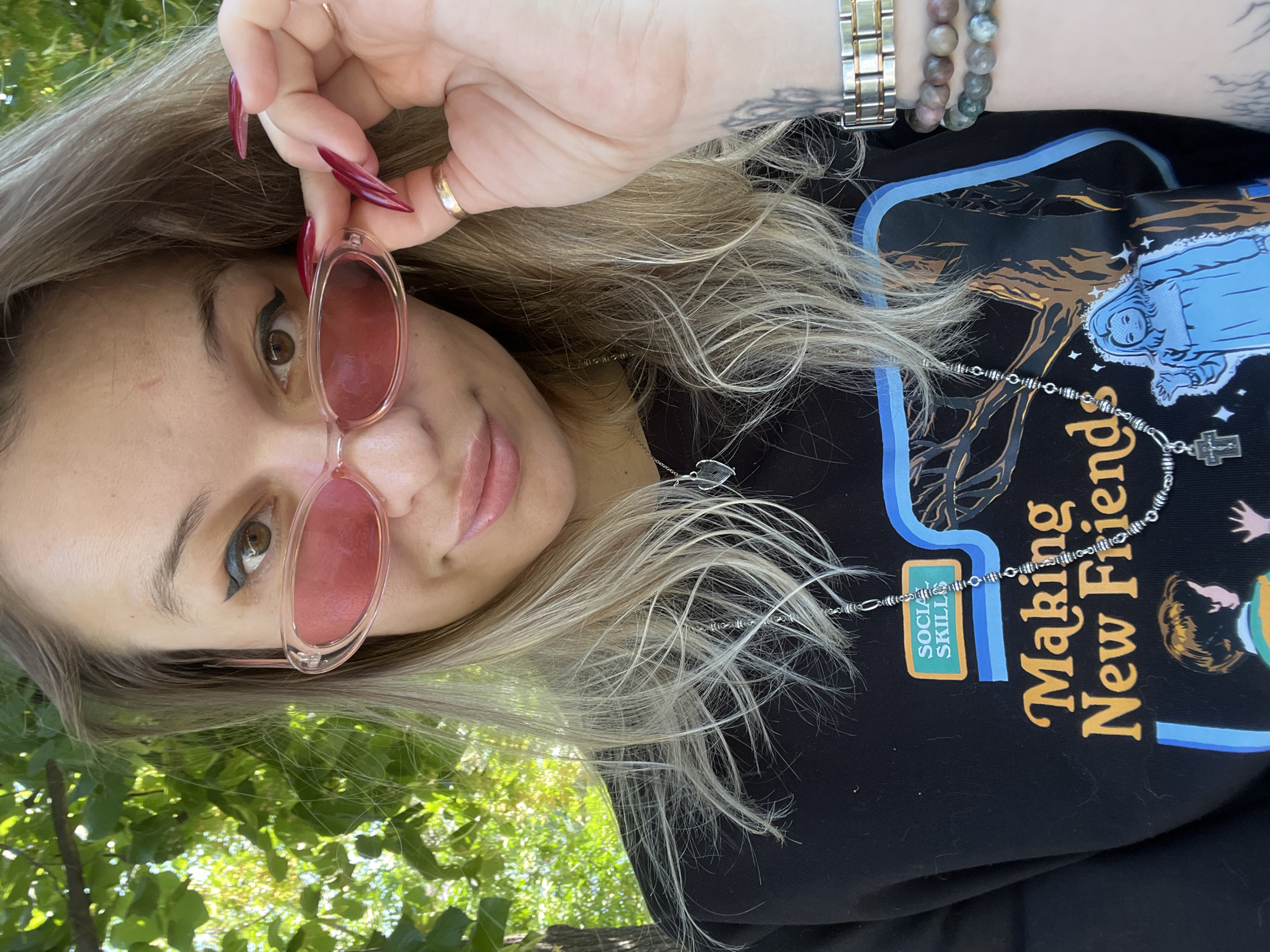 A medium shot captures a woman looking at the camera outdoors, partially obscured by tree foliage. She is wearing pink oval-shaped sunglasses and has long, blonde, wavy hair. Her right hand, adorned with long, dark red acrylic nails, is raised to adjust her sunglasses, revealing a gold ring and a silver watch with gold accents. She is also wearing a black crewneck sweatshirt with a graphic print. The graphic features the text "Making New Friends" in yellow and orange, with an illustration of what appears to be a doll or a young girl in a blue dress. The background shows blurred green foliage and tree trunks.