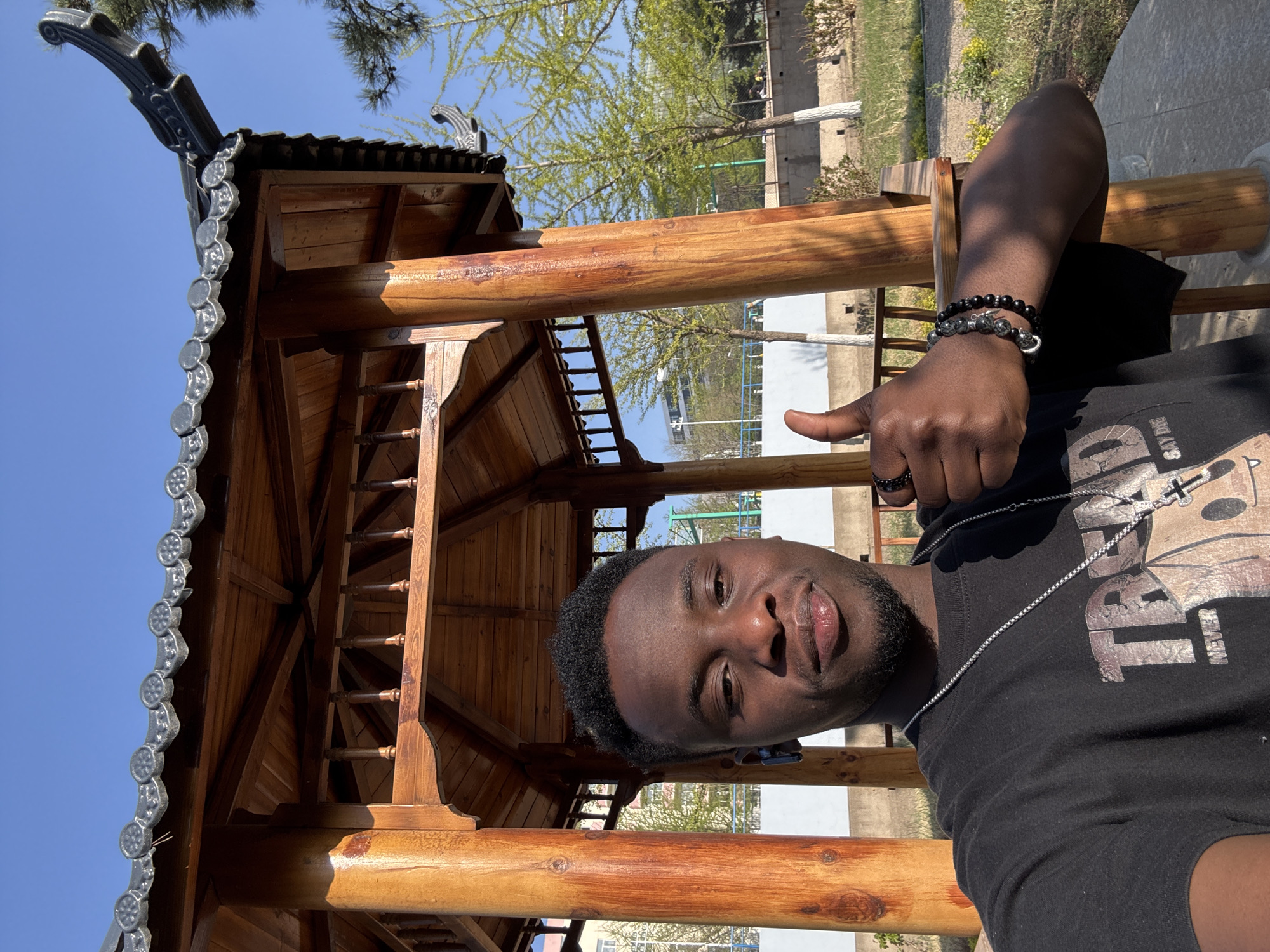 A young man with dark skin and a short beard takes a selfie within a traditional wooden gazebo under a clear blue sky, likely in Zhongtun, China. He gives a "thumbs up" gesture with his right hand, expressing a positive sentiment. He is wearing a black t-shirt, a silver chain necklace, black and grey beaded bracelets on his left wrist, and a black ring on his left ring finger. The black t-shirt features a silver graphic, with the word "NEVER" visible vertically on the right side of a larger, stylized metallic design.
The gazebo is constructed from polished light-brown wood, featuring sturdy beams, intricate joinery, and a decorative roof with dark, ornate tiles. The roof tiles have a distinctive scalloped edge with circular patterns and curved, sculptural end pieces. The overall architecture suggests a traditional Chinese design, typical of a park or garden setting.
The background shows vibrant green foliage on trees, indicative of spring, under a bright, sunny sky. The strong sunlight casts clear shadows, suggesting it is midday or early afternoon. Distant elements like fences or buildings can be faintly seen through the trees. The man's expression is neutral to slightly smiling, reflecting the casual and positive nature of the selfie.