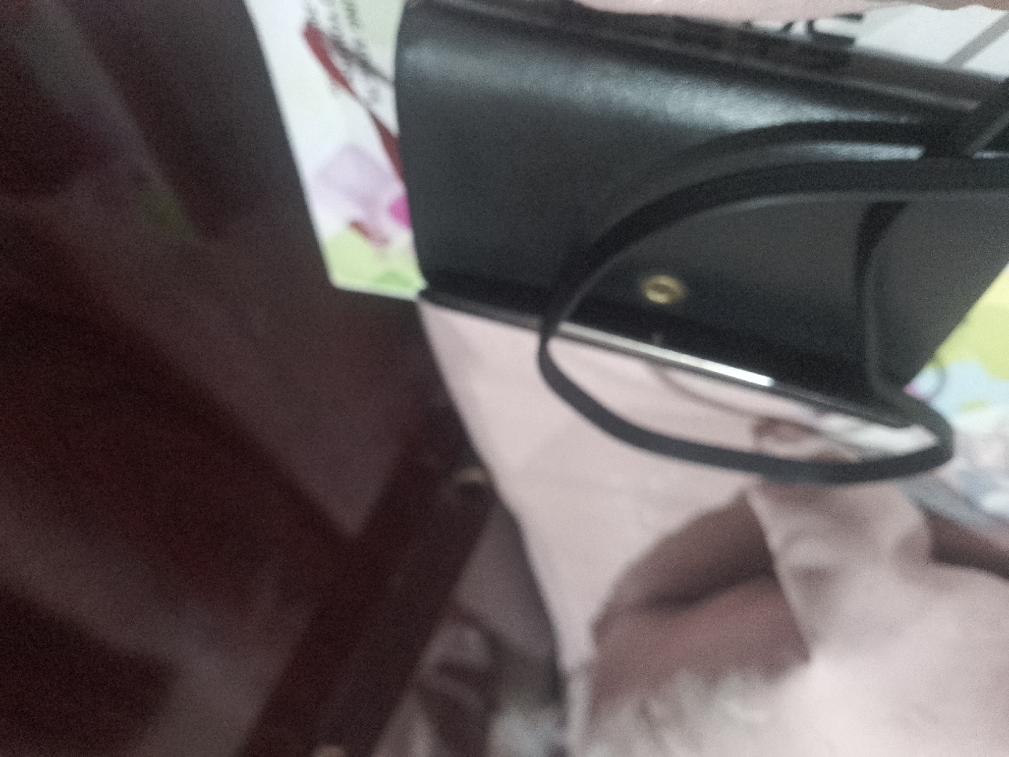 The image shows a close-up, angled shot of a black wallet resting on a pink surface. A thin black strap is visible, attached to the wallet and looping over the top. A small, circular metallic accent is also visible on the wallet. The background is dark and out of focus, with hints of what appears to be a wooden surface on the left. The lighting is dim and diffused. The location is unknown.