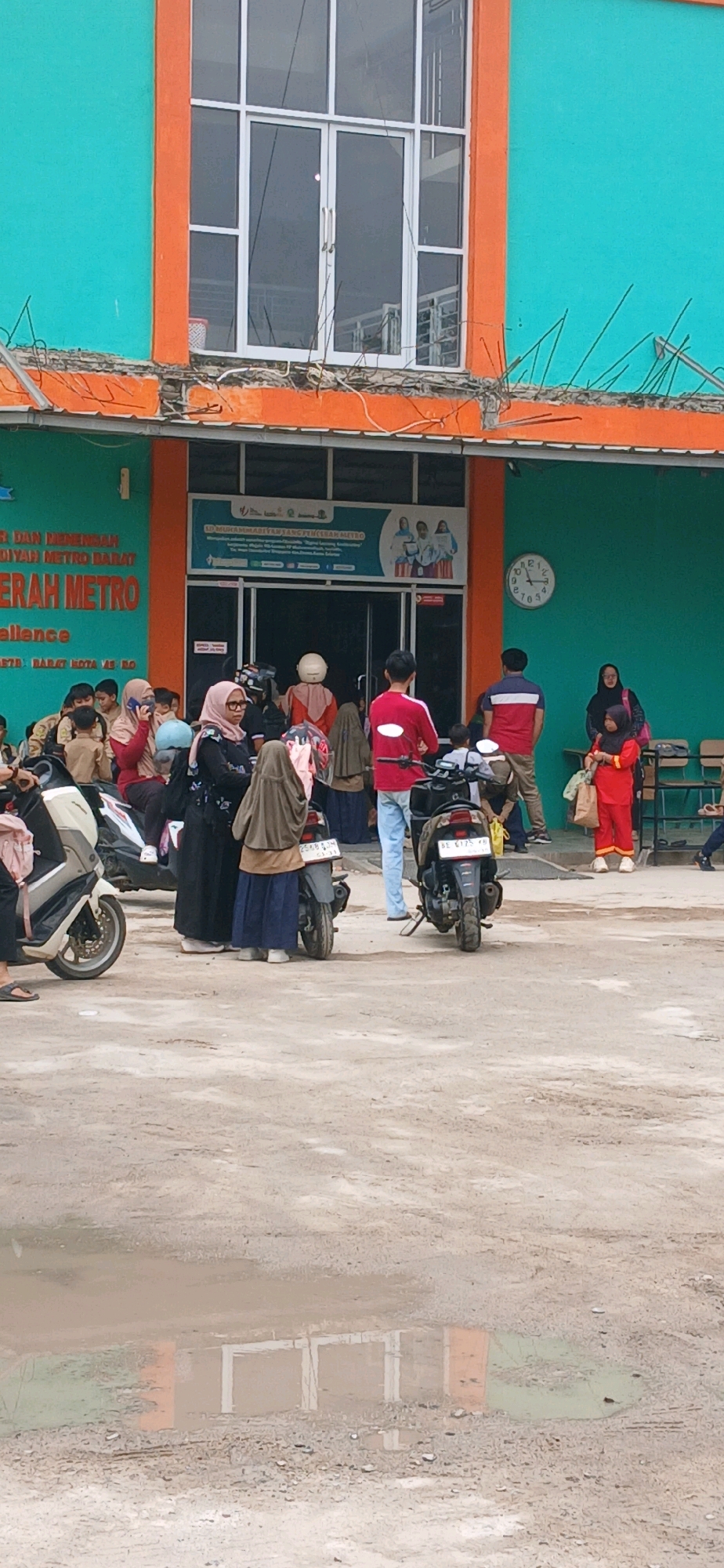 The image depicts an outdoor scene in front of a building with vibrant turquoise and orange accents. The building features large windows and an entrance with a sign that appears to be in Indonesian, suggesting a location in Indonesia. Specifically, the sign visible on the building reads "SD MUHAMMADIYAH METRO BARAT" and "SMK METRO", indicating that this is a school complex in the Metro district of West Jakarta.
In front of the building, there is a group of people, many of whom are wearing hijabs, suggesting a predominantly Muslim population. Several individuals are standing around, with some appearing to be waiting or conversing. There are also motorcycles parked in the foreground, typical for transportation in the region. The ground is a mix of dirt and puddles, indicating recent rain or a damp environment. A large clock is visible on the turquoise wall of the building, displaying the time as approximately 10:08. The overall atmosphere appears to be a busy, everyday scene in front of an educational institution.