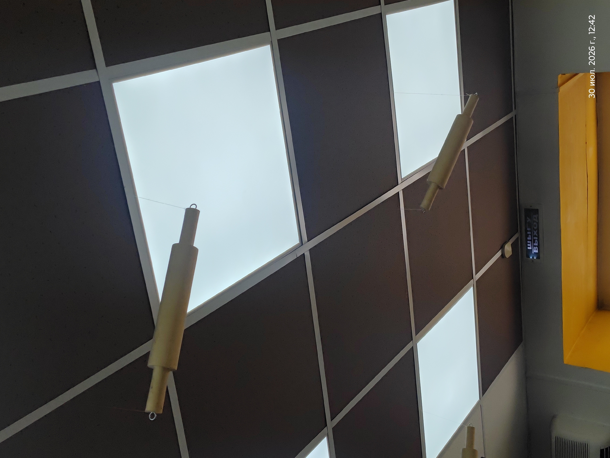 The image captures a view looking up at a suspended ceiling with large, square, illuminated panels and dark brown square acoustic tiles. Several rolling pin-shaped objects are hanging from thin wires, serving as decorative elements. In the lower part of the image, a sign displays "ВЫХОД" (EXIT) in Cyrillic, illuminated in blue. Below this sign, a bright yellow architectural element is visible, possibly part of a doorway or counter. The timestamp "30 июл. 2026 г., 12:42" is superimposed in the lower right corner, indicating the date and time the photograph was taken. The location is identified as Astana, Kazakhstan.