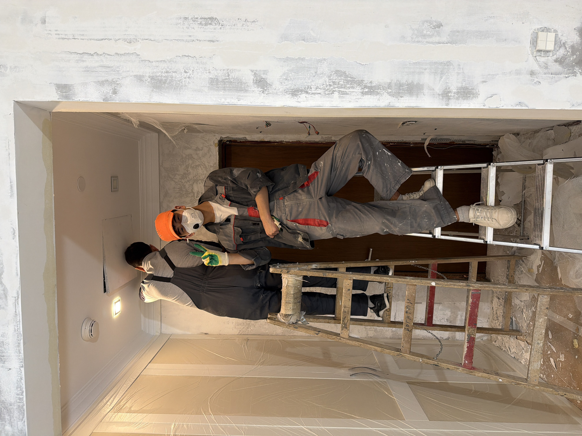Two men are working on what appears to be a renovation or repair project inside a building. The men are dressed in work clothing, including overalls, and one is wearing a hard hat and a face mask. They are standing on ladders of two distinct types to reach the ceiling area. The surroundings indicate an interior space under construction, with a partially finished ceiling, exposed walls, and protective plastic sheeting covering the floor. The location appears to be in Khimki, Russia.