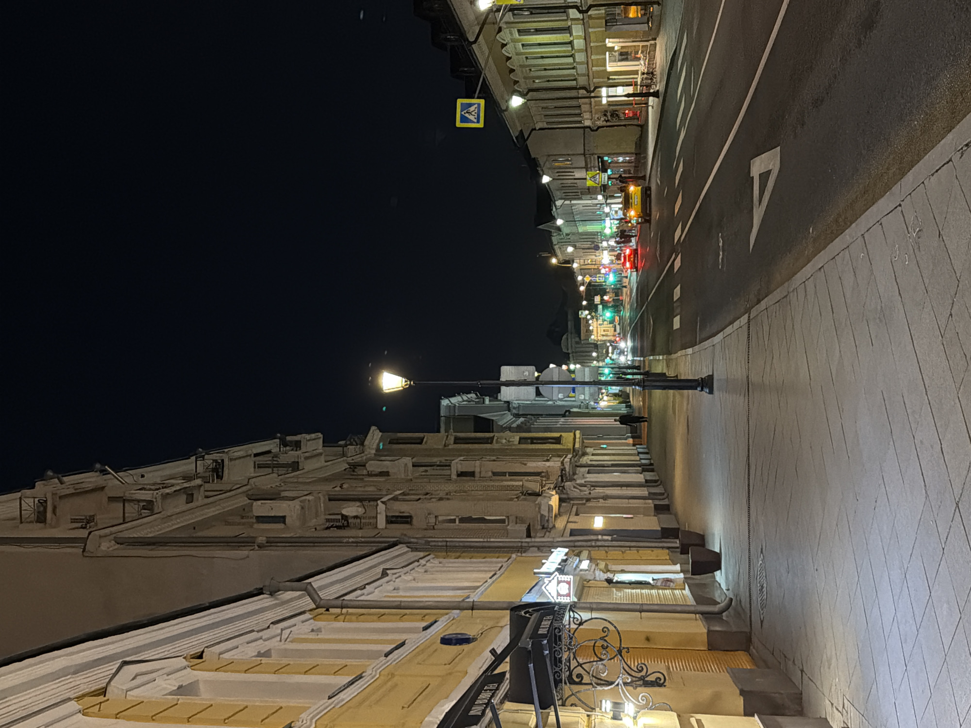 The image depicts a street scene at night in Moscow, Russia. The perspective is from a sidewalk on the left side of the street, looking down the length of the road. On the left, a yellow building with ornate architectural details and balconies runs along the sidewalk. Shops with awnings are visible at street level. A traditional streetlamp illuminates the sidewalk.
The street itself is paved with asphalt and has white lane markings and a pedestrian crossing. Several cars are visible on the road, including a yellow taxi and a red car, their headlights and taillights creating streaks of light. Traffic lights are visible in the distance, with some showing green. Pedestrians are sparsely distributed along the sidewalk and near the road.
On the right side of the street, more buildings line the road, also featuring traditional architecture and illuminated windows. A pedestrian crossing sign is mounted on a pole. The overall atmosphere is that of a quiet urban evening. The sky is dark, indicating nighttime.
