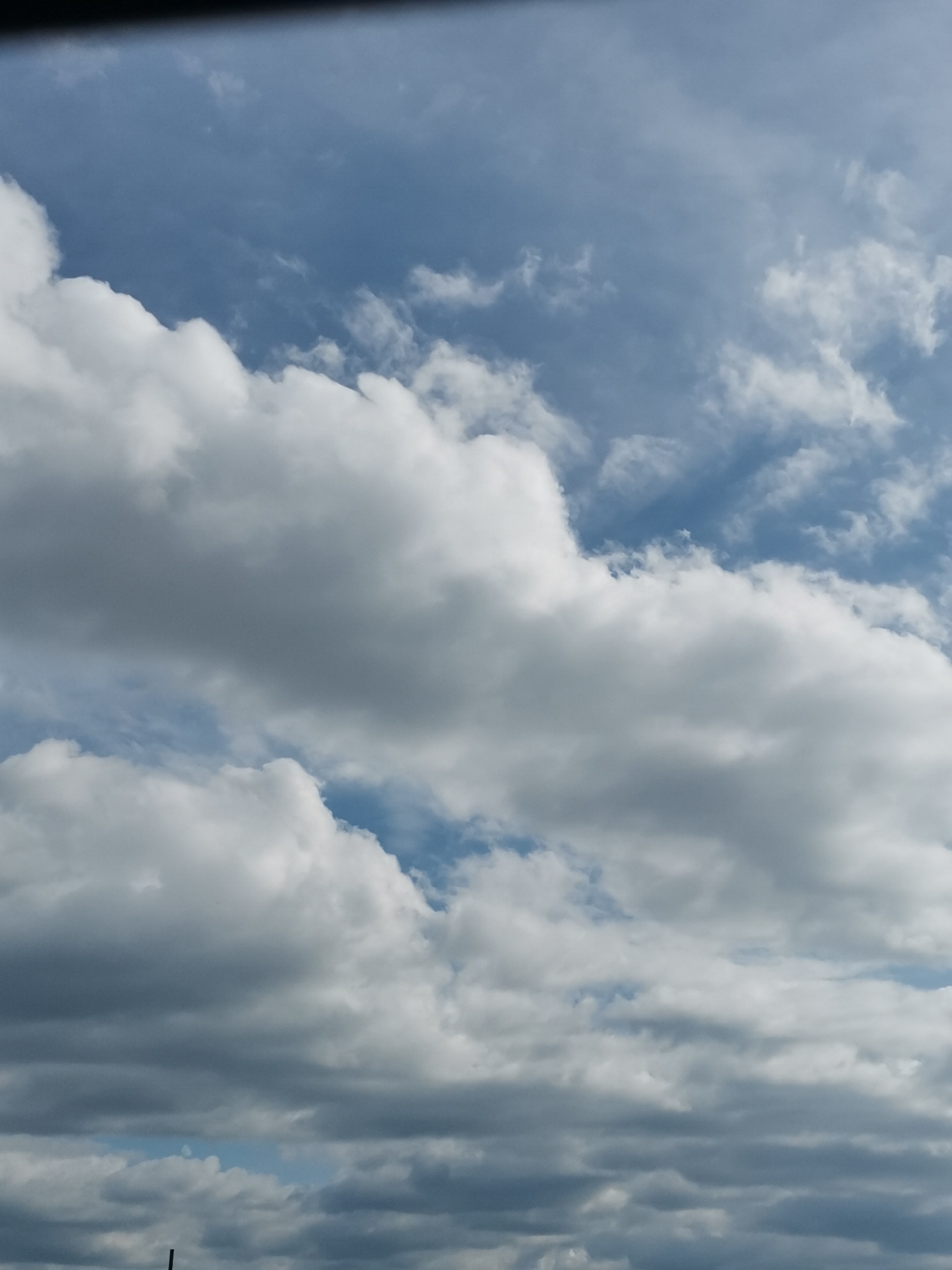 The image shows a view of the sky filled with clouds, with a dark band across the top, possibly indicating the edge of a roof or structure. The sky is a mixture of blues and grays, with dense white cumulus clouds dominating the foreground and midground, some of which appear shadowed. There are also thinner, wispy clouds present. A small sliver of blue sky is visible in the upper right portion of the frame. The lower portion of the image shows a darker, more uniform layer of clouds. The scene is a natural skyscape, not specific to any particular location in Sofia, Bulgaria, as there are no discernible landmarks or urban elements.