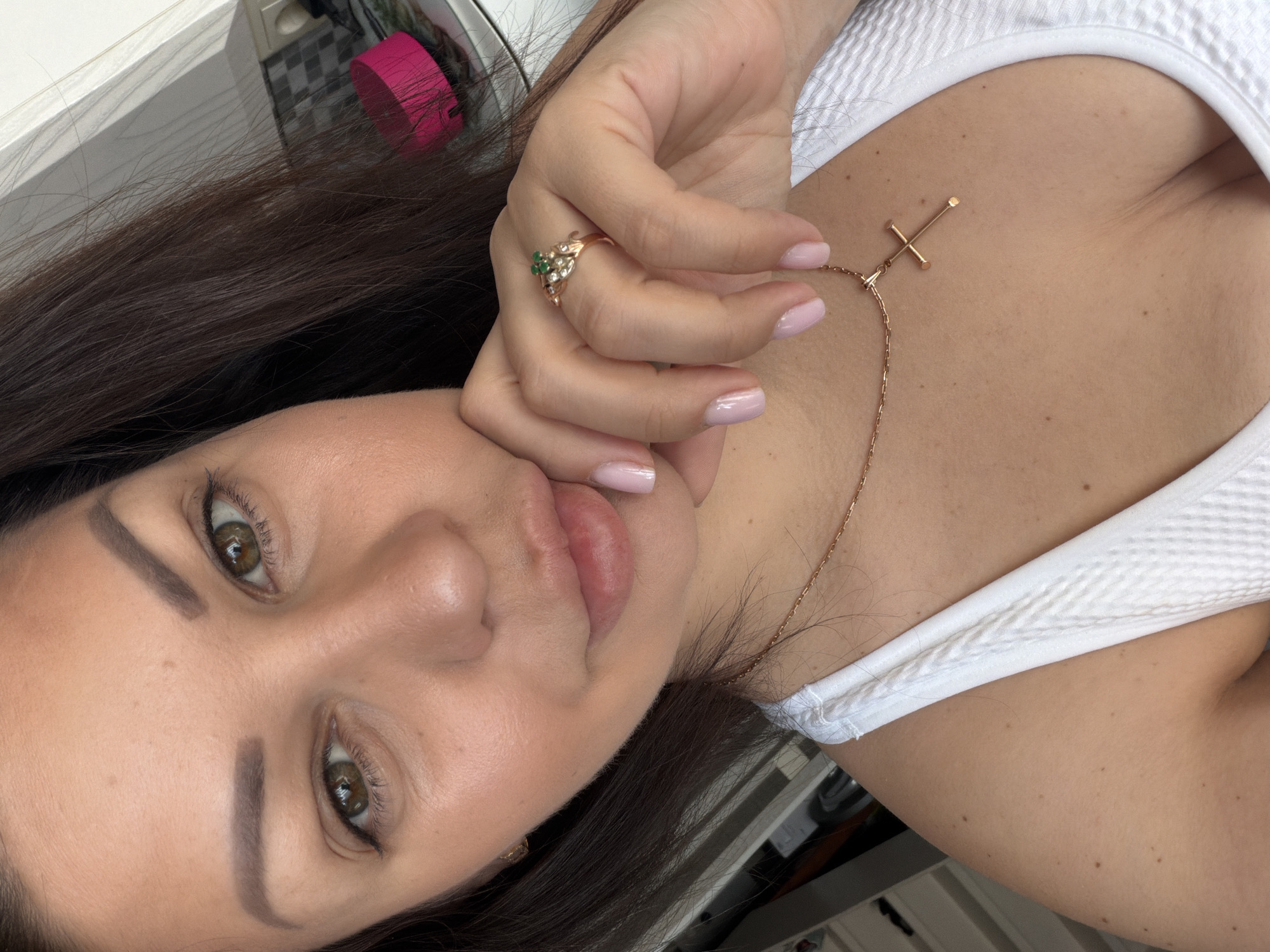 A close-up selfie captures an adult female indoors in Sochi, Russia. She has long, dark brown hair, light-to-medium skin tone, and hazel eyes. Her eyebrows are well-defined, and her lips appear full. She is wearing a white ribbed or textured tank top, revealing partial cleavage. Around her neck is a gold-toned necklace with a minimalist cross pendant. On the ring finger of her right hand (viewer's left), she wears a gold-toned ring featuring a cluster of small green stones, possibly emeralds, surrounded by smaller clear stones. Her fingernails are manicured with light pink polish. Her right hand is positioned with fingers curled, resting against her jawline. She looks directly into the camera with a neutral to slightly smiling expression. The background elements indicate an interior space, likely a room with white walls or cabinetry. Visible on the right side of the frame (viewer's left) are hints of a light-colored surface and a bright pink cylindrical object.