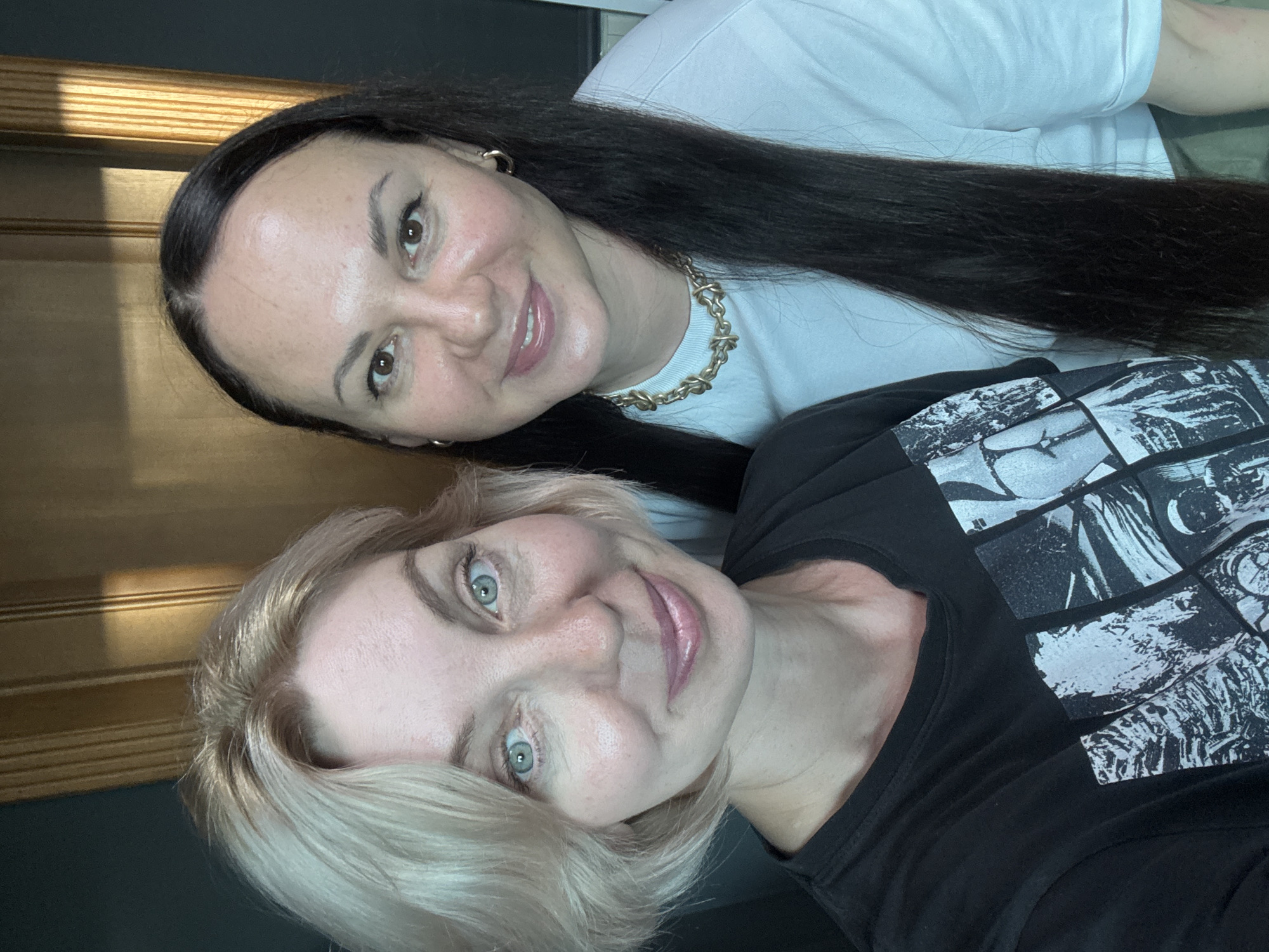 Two women are posing for a selfie in front of a wooden door. The woman on the left has short, blonde hair and is wearing a black t-shirt with a graphic print. She is smiling and looking at the camera with blue-green eyes. The woman on the right has long, dark hair and is wearing a white t-shirt and a gold chain necklace. She is also smiling and looking at the camera. The background appears to be indoors, with a dark wall and a wooden door with vertical slats. The lighting suggests it may be daytime. The image quality is good, with clear details of the subjects and their surroundings. There is no identifiable information about the location within the city of Sochi, Russia, or any other specific context.