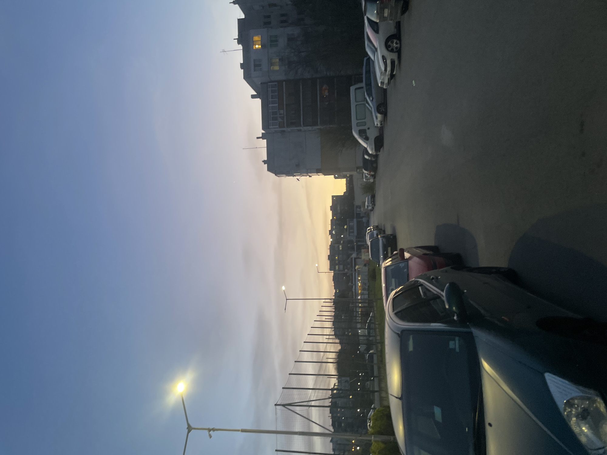 This image captures an urban street scene in Razgrad, Bulgaria, during the transition from daylight to dusk. The sky dominates the upper portion, showcasing a gradient from deep blue on the left to soft orange and yellow hues on the right, indicative of a sunset or twilight.
In the mid-ground, a line of multi-story residential buildings is visible, characterized by their somewhat grey and worn facades. Several windows in these buildings are lit, casting warm, yellow glows that stand out against the fading natural light. Below the buildings, a street or parking area stretches, populated by numerous parked cars. In the immediate foreground on the right, a dark blue or green sedan is prominently featured, alongside other vehicles including a white van and a silver car further down the street.
Two streetlights are visible: one in the lower left corner brightly illuminates its surroundings with a strong yellow glow, and another can be seen further in the distance. A tall, dark fence or netting, possibly enclosing a sports field, runs horizontally across the middle distance of the image, separating the parked cars from the buildings and the horizon.
No people are visible, and no specific activity is occurring, lending a quiet and still atmosphere to the scene. The artificial lights from the streetlamps and building windows contrast with the fading natural light, highlighting the arrival of evening. There is no visible text in the image.