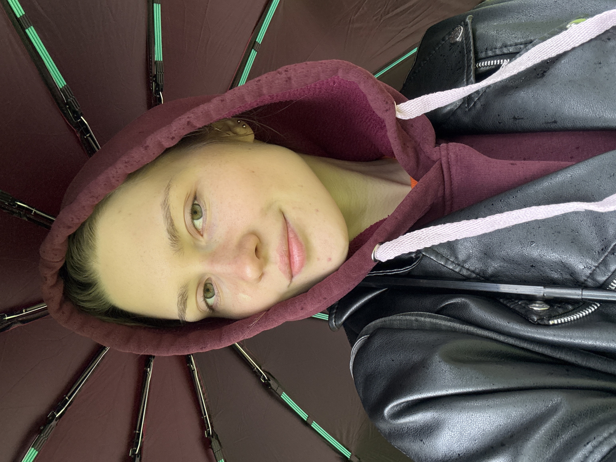 A young person with light skin and light hair is under a dark red umbrella, wearing a burgundy hooded sweatshirt and a black leather jacket. The individual is looking at the camera with a slight smile. The umbrella has green accents on its supporting structure. The background is not visible, so the location cannot be determined. There is no evidence to suggest this is in Saint Petersburg, Russia.