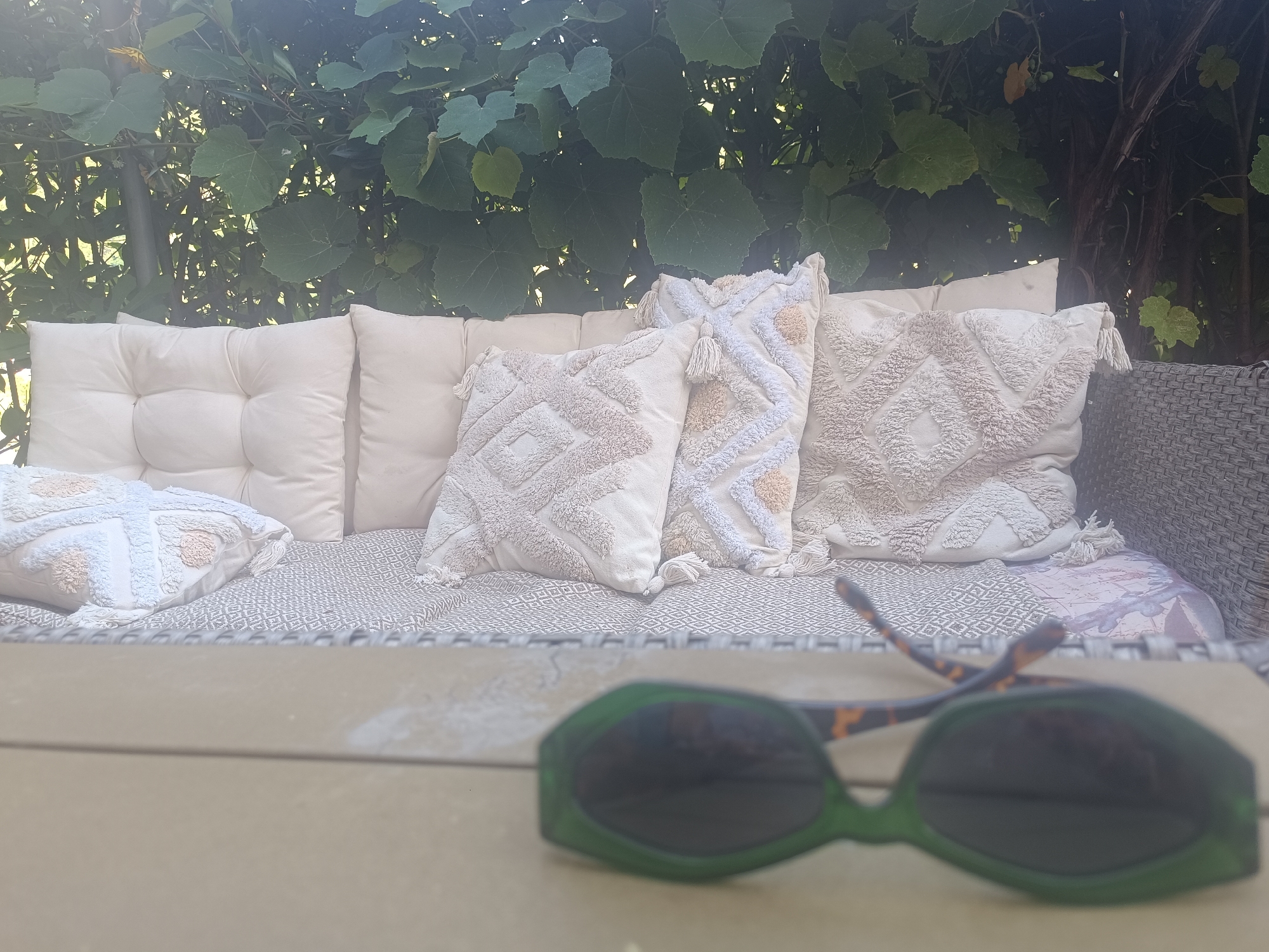 This is a medium-shot, eye-level, outdoor photograph of a patio area with a wicker-style sofa adorned with several textured, neutral-colored cushions, including one with a diamond pattern and tassels, and another with a cross-stitch pattern. The sofa is set against a backdrop of lush green foliage, likely from a vine or climbing plant, with some sunlight filtering through. In the foreground, a pair of green-tinted sunglasses with tortoise-shell arms rests on a light-colored wooden surface, possibly a table. The scene is tranquil and appears to be a comfortable outdoor seating area. The location is identified as Serravalle, Switzerland.