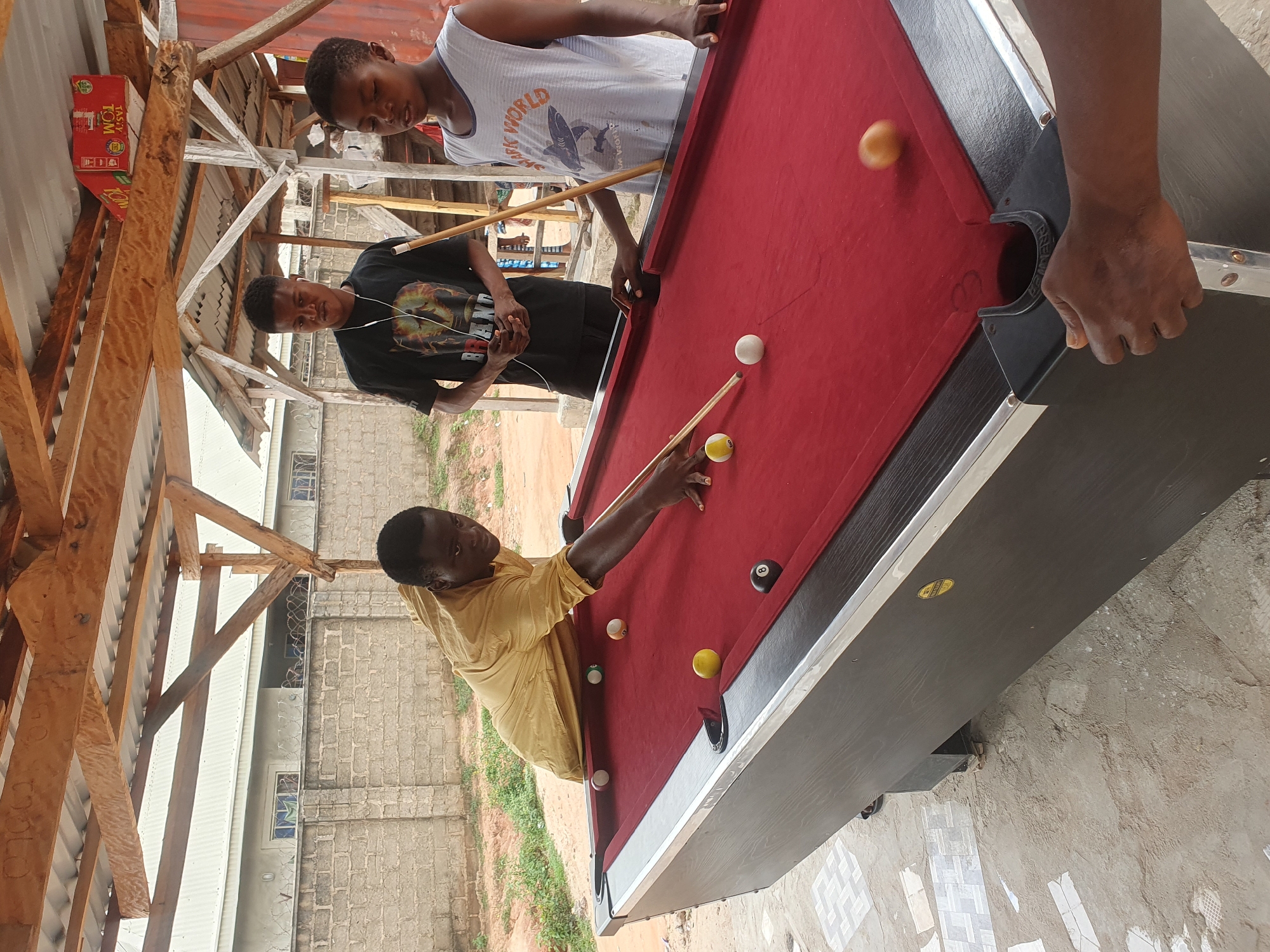 In Lokoja, Nigeria, three young males are engaged in a game of pool under a wooden structure with a corrugated iron roof. One individual, wearing a yellow tunic, is actively playing, leaning over the red felt pool table, cue in hand, preparing to strike a ball. Another male, dressed in a black t-shirt with a graphic print and wearing white earphones, stands behind the table, observing the game. The third young male, in a white tank top with a shark graphic, also watches the game. The pool table has several balls on its surface, including a black 8-ball. The environment appears to be an outdoor space with a brick wall and some vegetation visible in the background. The ground is a mix of concrete and dirt.