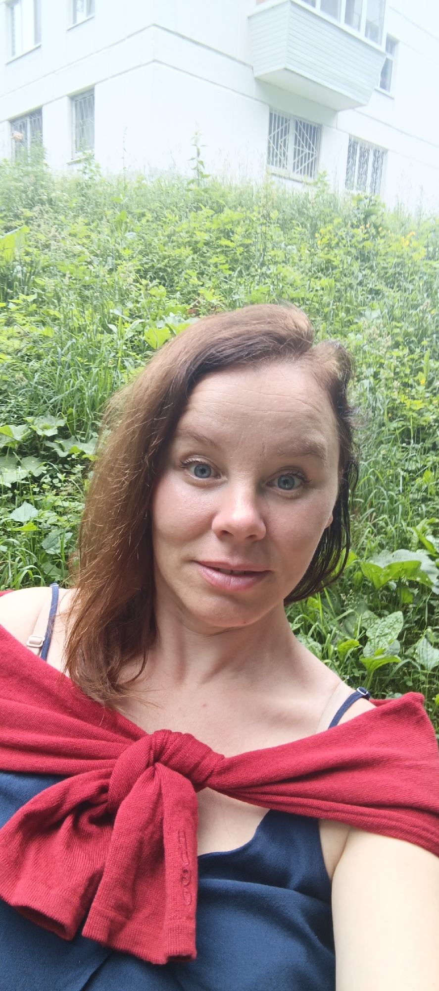 A woman with shoulder-length brown hair and blue eyes is pictured in a selfie. She is wearing a blue top and a red sweater tied around her shoulders. The background consists of lush green foliage and a white building with windows, possibly a multi-story apartment building. The setting appears to be outdoors in a residential area, potentially in Chaikovsky, Russia, given the architectural style of the building and the vegetation.