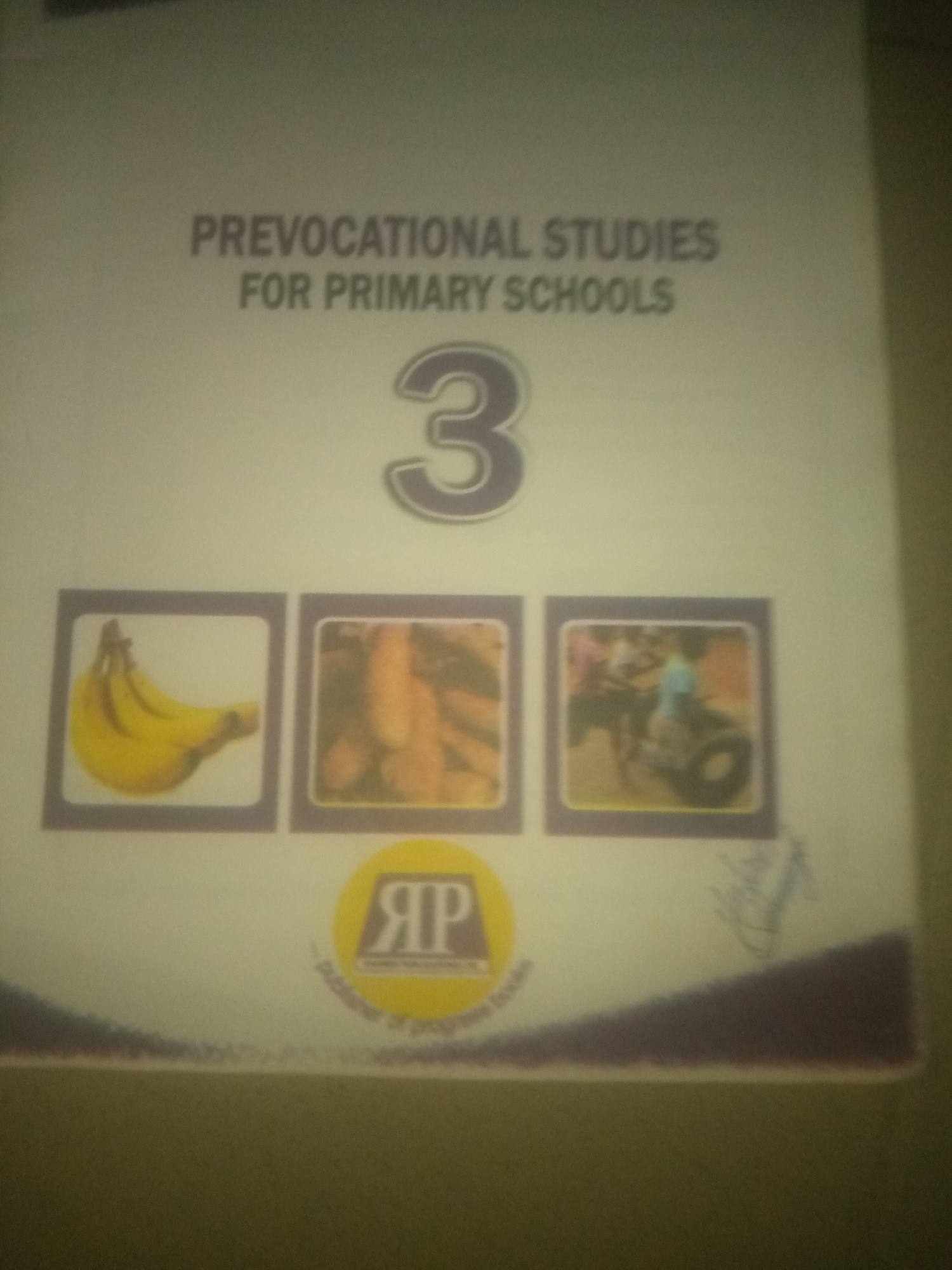 The image displays the cover of a book titled "PREVOCATIONAL STUDIES FOR PRIMARY SCHOOLS 3". The cover features illustrations of bananas, carrots, and a person riding a bicycle. A logo with the letters "RP" is also visible. The overall presentation is that of an educational book, likely for children. There are no discernible elements within the image that would indicate a specific location in the city of Jauro Yinu, Nigeria. The image is a close-up of the book cover and does not provide any environmental context.