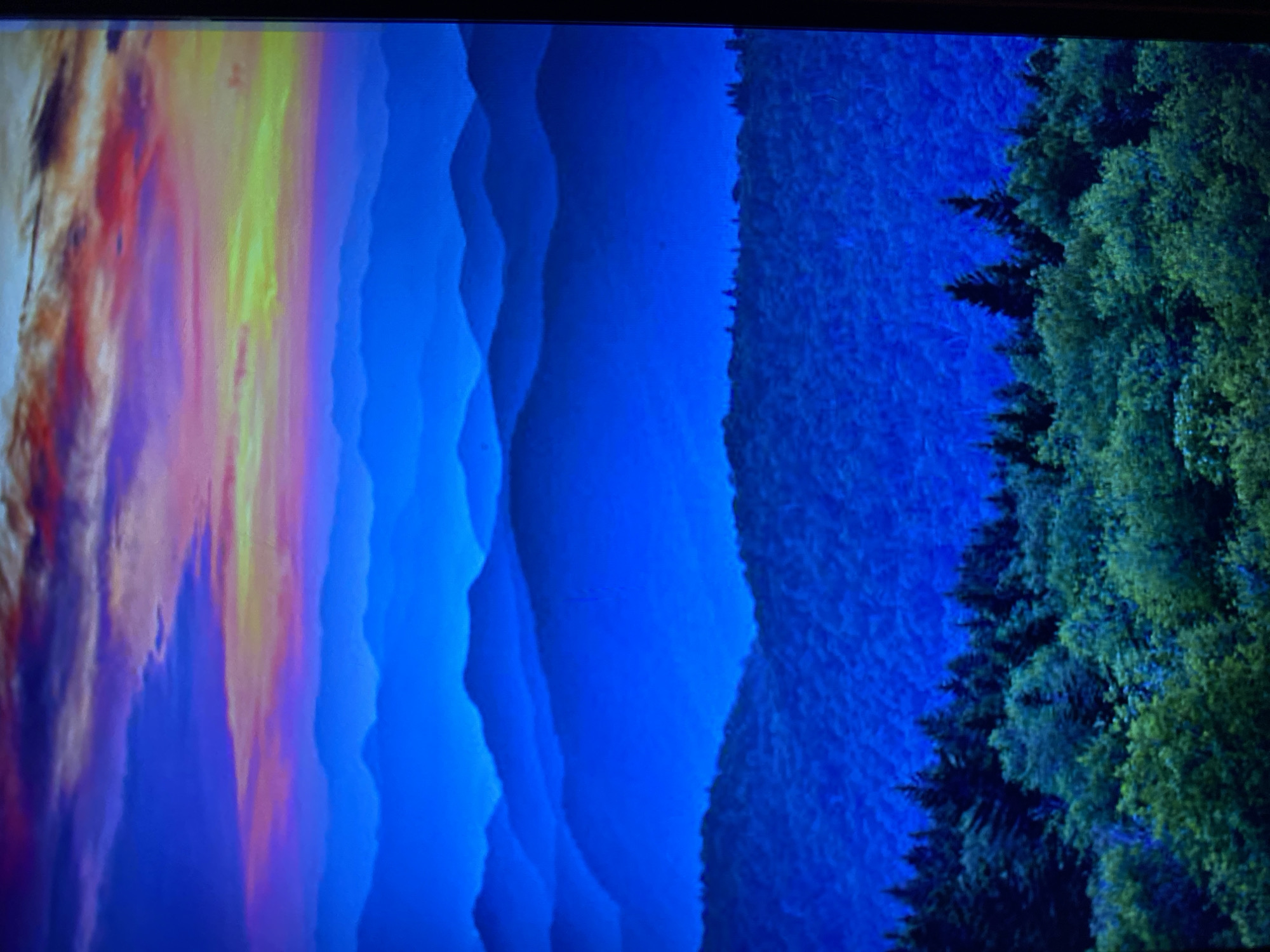 The image displays a panoramic view of layered mountain ranges under a vibrant, colorful sky. The foreground features a dense forest with lush green trees. The mid-ground consists of rolling hills and mountains covered in foliage, gradually fading into darker blue hues with distance. The background is dominated by successive mountain silhouettes, each appearing lighter and more indistinct than the one in front, creating a sense of depth. The sky above is a striking gradient of blues, purples, and oranges, indicative of a sunset or sunrise, with scattered clouds. The scene is a natural landscape, and there is no indication of any urban environment or specific location in Luh, Ukraine. There are no people or discernible objects other than the natural elements of trees, mountains, and sky.