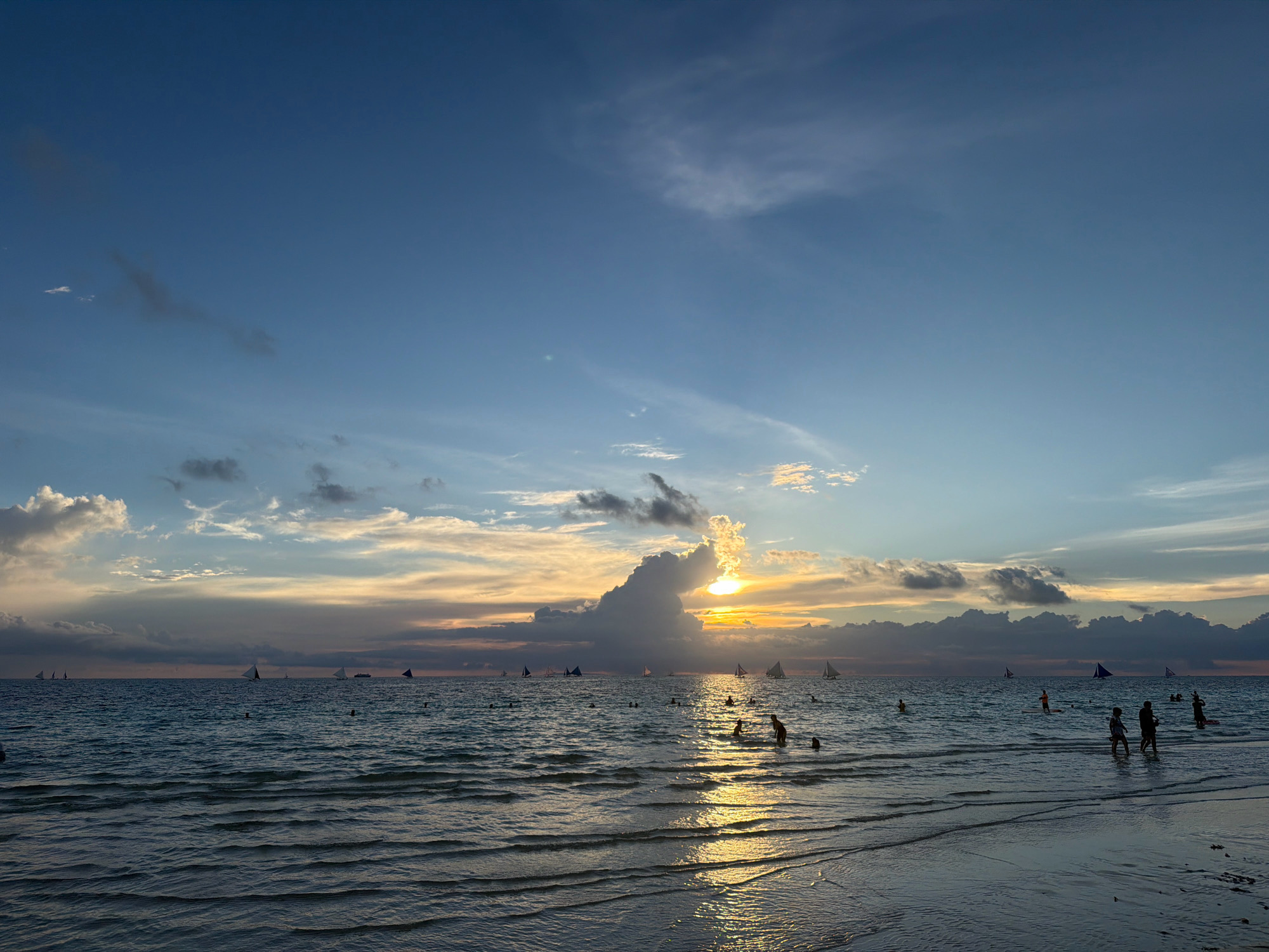 The image depicts a sunset scene at White Beach in Malay, Philippines. The sky is a gradient of blue, with scattered clouds that are tinged with orange and yellow from the setting sun. The sun itself is partially obscured by a large, dark cloud formation, creating a dramatic effect as its rays pierce through. The ocean is calm, with gentle waves creating rippling reflections of the sky and the setting sun on its surface. Numerous sailboats with white sails are visible on the horizon, adding to the serene atmosphere. In the foreground, several people are wading in the shallow waters near the sandy shore. The overall impression is one of a peaceful and beautiful tropical evening.