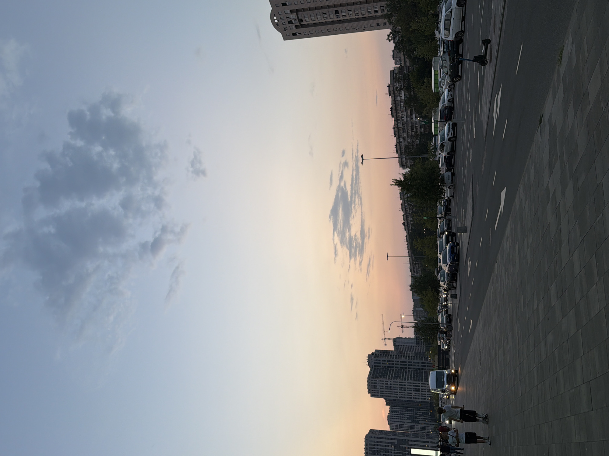 The image captures a street scene in Kyiv, Ukraine, during what appears to be dusk. Tall, modern apartment buildings and construction cranes dominate the skyline on the left side of the frame. A white van is driving on the road, with its headlights illuminated. Several cars are parked in a lot to the right of the road. Two individuals are walking on the paved sidewalk in the foreground. The sky is a soft gradient of pale blue and pink, with scattered clouds. The overall atmosphere is calm and urban.