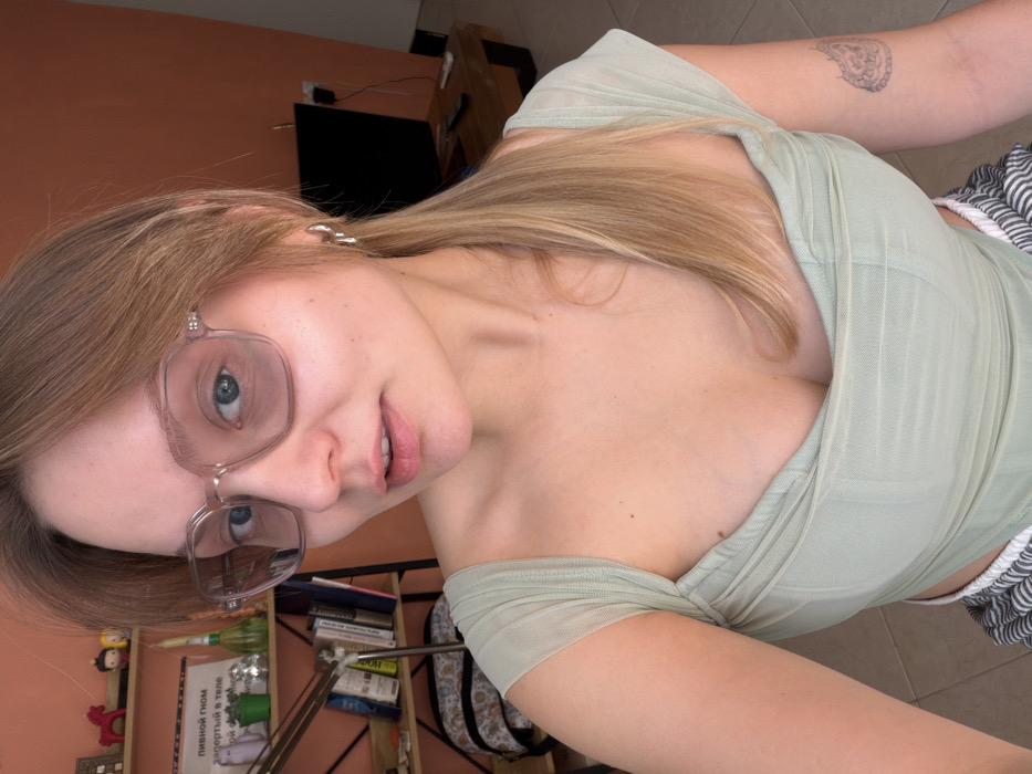 A fair-skinned female subject is depicted indoors in what is identified as a residential setting in Tel-Aviv, Israel. She is seen from the chest up, facing the camera with a direct gaze. Her long, blonde hair is styled in a ponytail, with some strands framing her face. She wears hexagonal-shaped, lightly tinted eyeglasses and light pink lipstick. A light green, sheer top with cap sleeves and a sweetheart neckline covers her torso. A small, light-colored tattoo with an intricate pattern is visible on her left bicep. Silver-colored hoop earrings with a dangle are present on her right ear. Striped bottoms are visible at the bottom edge of the frame.
The background on the left features a light orange wall with a wooden shelving unit. Objects on the shelves include a red horse figurine, small decorative figures, a potted plant, glass items, and a stack of books. A white paper sign with Cyrillic text, "ПИВНОЙ ГНОМ запертый в теле" (BEER GNOME trapped in the body), is affixed to the wall above the shelves. A dark-colored backpack with a patterned design rests below the shelves, next to a metal lamp stand.
The background on the right side of the image reveals a darker orange wall with a flat-screen television positioned on a low, dark wooden table. A white cable extends from behind the television. The floor, visible at the bottom of the frame, is covered in light-colored tiles. The scene is illuminated by natural light, suggesting daytime.