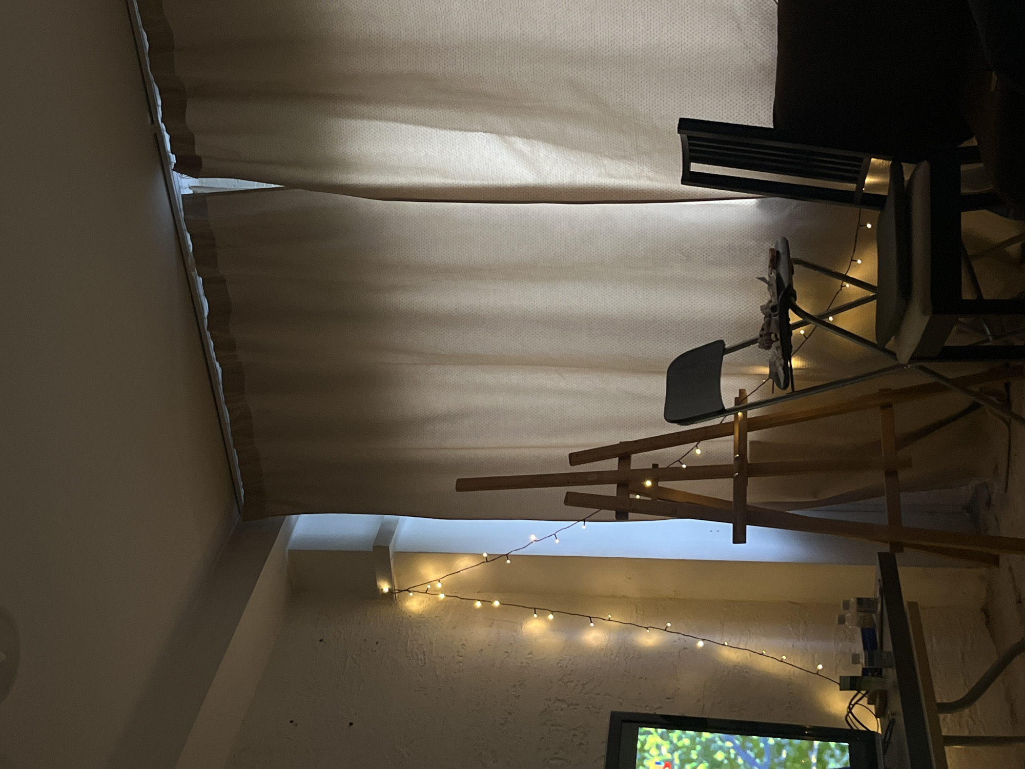The image shows an interior space with a low ceiling draped in a beige fabric. A chair with a wooden frame and a dark seat is positioned prominently in the scene. A string of lights is hung across the wall, and a television screen is visible at the bottom. A wooden structure, possibly a shelf or decorative element, is also present. The image's location and specific details suggestive of Krasnoyarsk, Russia, are not discernible.