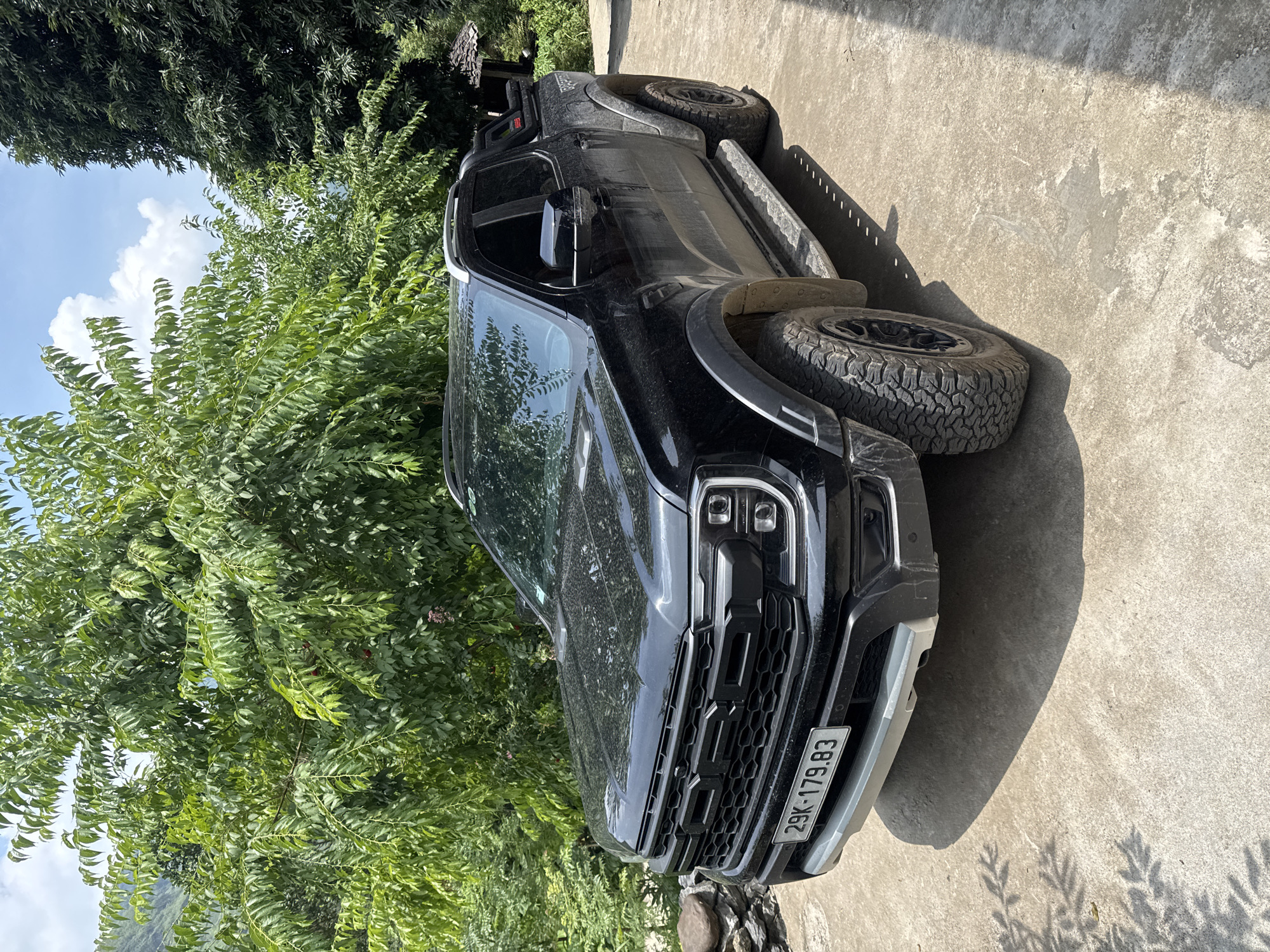 A black Ford Ranger pickup truck, likely a Raptor model given the prominent "FORD" lettering on the grille, is parked on a concrete surface. The truck is positioned at an angle, with its front facing slightly towards the right and its left side running parallel to dense, lush green trees and bushes that dominate the left side of the frame. The vehicle appears somewhat dusty, suggesting recent travel or outdoor exposure. It features robust all-terrain tires and side steps.
The scene is brightly lit by natural daylight, casting distinct shadows on the concrete from the truck and surrounding foliage, indicating a sunny day, likely during the mid-morning or afternoon. Patches of blue sky with white clouds are visible above the treetops. The greenery is vibrant and dense, creating a natural backdrop.
Visible on the front of the truck is a license plate displaying the text "29K-179.83". The overall setting evokes an outdoor, possibly rural or semi-rural, environment consistent with the Du Già Commune in Vietnam. There are no people visible in the image, and the truck is stationary.