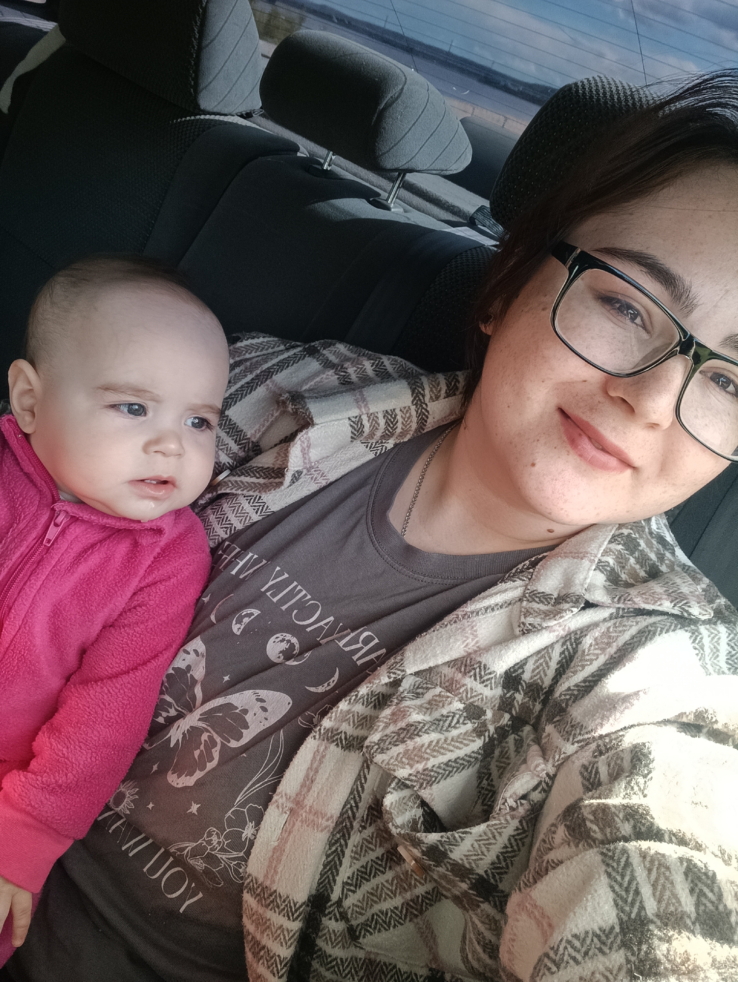A selfie taken inside a car shows a woman and a baby. The woman is wearing glasses, a grey t-shirt with a butterfly and moon phases design, and a white and brown plaid shirt. She has a silver chain around her neck. The baby is wearing a pink zip-up onesie. The background shows the interior of a car, including the headrest and the rear window. It's impossible to determine the exact location in the city of Chaikovsky, Russia, based solely on this image.