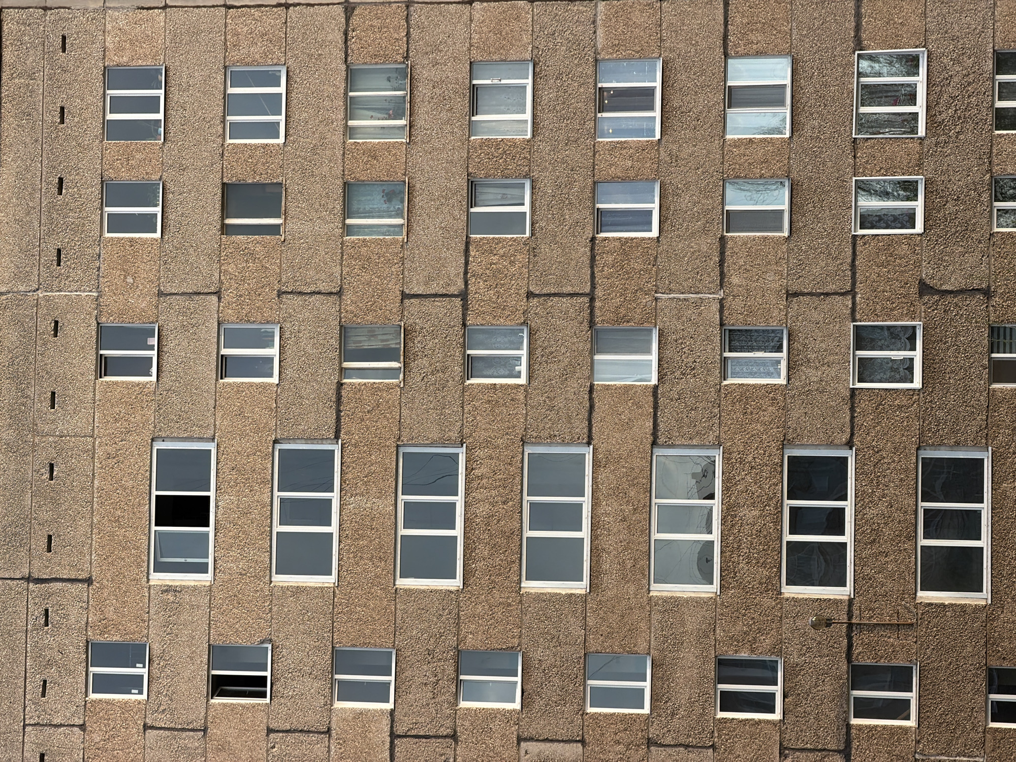 The image shows the facade of a multi-story building. The building appears to be constructed of pre-cast concrete panels with a rough, textured surface. The panels are a tan color. The building has numerous rectangular windows with white frames. The windows are arranged in a grid pattern across the facade. Some windows reflect the sky or interior lighting, while others appear dark. The overall impression is of a utilitarian and functional architectural style, potentially located in an urban environment in Izhevsk, Russia.