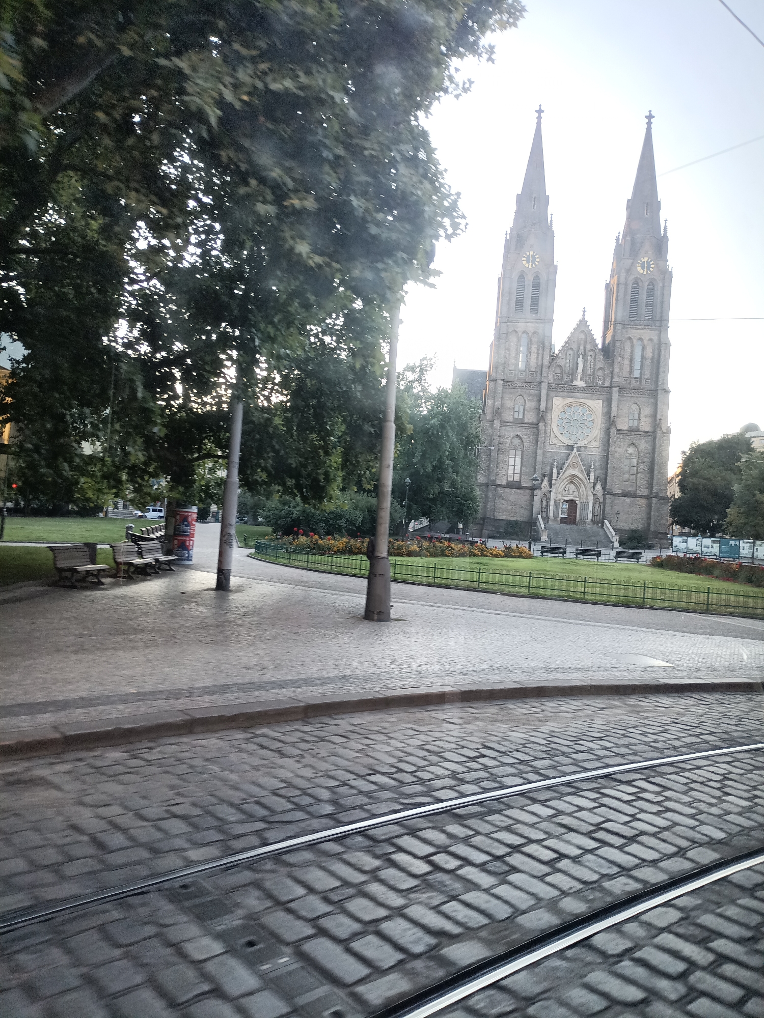 This is a photograph taken from a tram in Prague, Czechia, capturing the Basilica of St. Peter and St. Paul in Vyšehrad. The foreground shows tram tracks on a cobblestone street, with a slight motion blur indicating the tram is moving. To the left of the tracks is a paved pedestrian area with several wooden benches and a metal pole. Beyond the benches is a grassy area with trees and flowerbeds. The dominant feature of the background is the neo-Gothic Basilica of St. Peter and St. Paul, characterized by its twin spires, clocks, and intricate facade. The sky is bright and overcast, suggesting it is daytime. The overall scene depicts a public urban space with architectural landmarks and elements of urban transport.