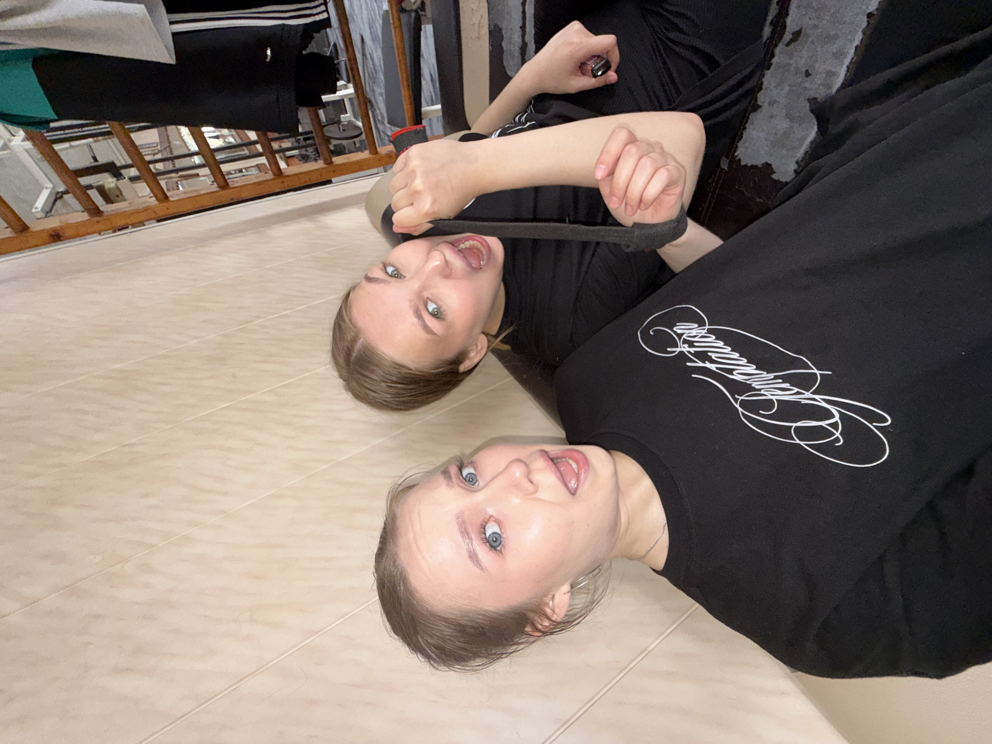 Two young women, likely identical or very similar in appearance, lie on a light-colored tiled floor, looking upwards towards the camera from an oblique angle. Both have light brown hair, light-colored eyes, and light complexions, and are wearing black long-sleeved tops.
The woman positioned further back playfully pulls a black strap or band towards her open mouth, with an expressive, energetic gaze directed at the camera. In her left hand, she holds a small, shiny, cylindrical object. The woman in the foreground also looks directly at the camera with a wide-eyed, open-mouthed, playful expression. On her black top, a white cursive inscription is partially visible, resembling "Simplicity" or a similar word.
The setting appears to be indoors, possibly a room in a residence, with a wooden railing structure visible in the background, which might be part of a bed frame or furniture. Some fabric or clothing hangs over this railing. A dark, textured surface is also present behind the background woman. The tiled floor is a light beige or cream color. The image captures a playful moment, with both subjects engaging directly with the camera. The time of day is not discernible, and while the location context provided is Chaikovsky, Russia, there are no specific visual cues in the image to confirm this.
