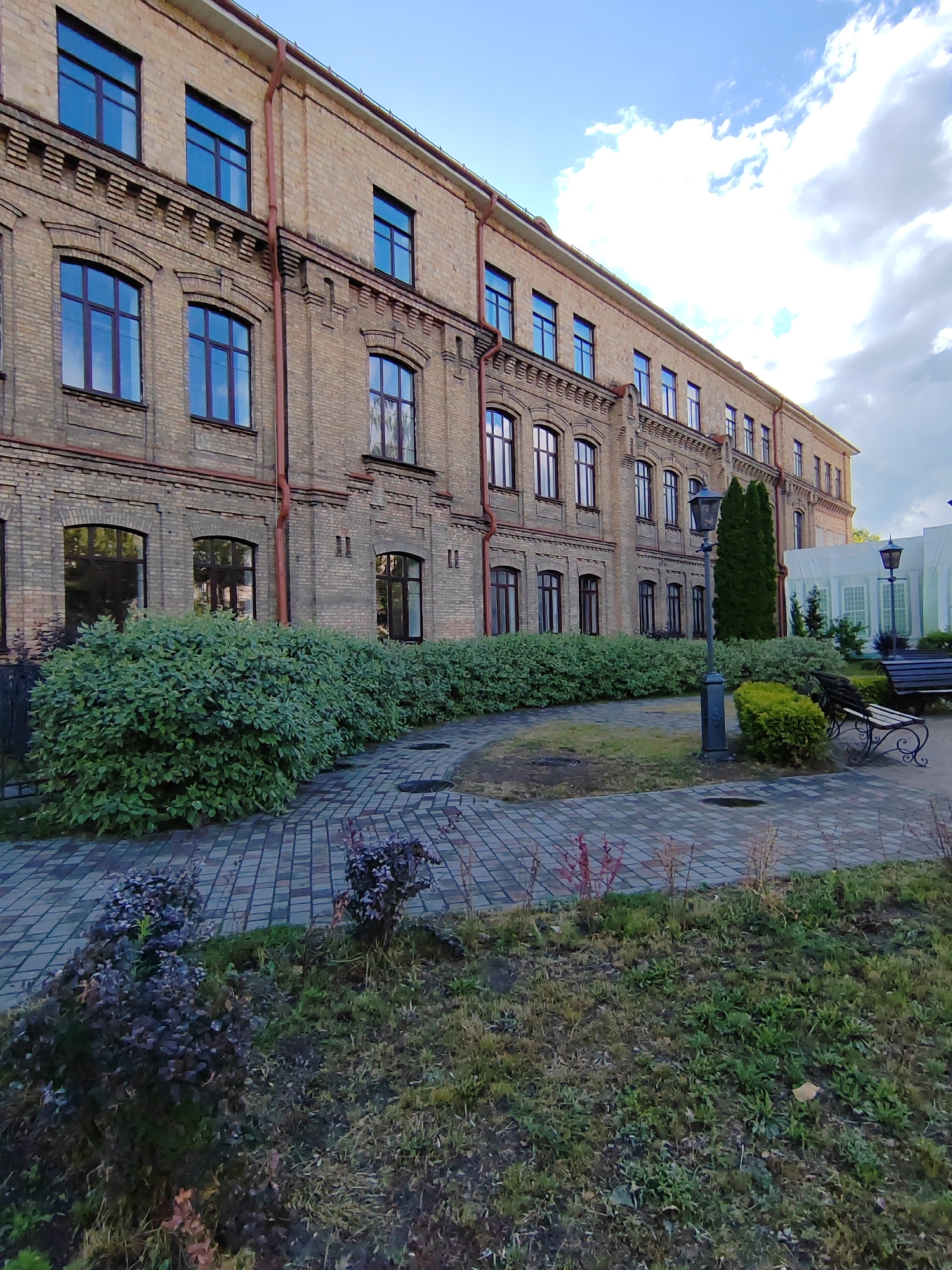 The image shows a brick building with many windows, a sidewalk, and some greenery. The building is several stories tall and appears to be quite old, with ornate brickwork and a red gutter running along the side. The windows are tall and rectangular, with dark frames. The sidewalk is paved with interlocking bricks, and there is a line of dense green bushes running along the side of the building. In the foreground, there are some smaller bushes and patches of grass. There are also two lampposts and a bench visible in the scene. The sky is partly cloudy, with a mix of blue sky and white clouds.