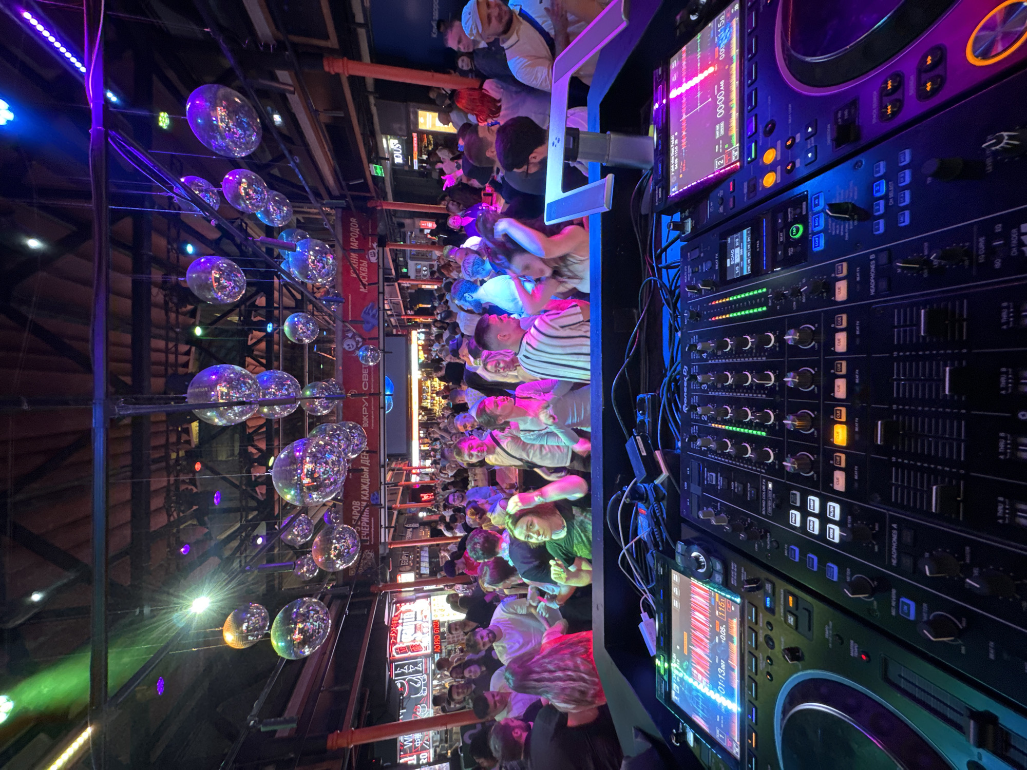 The image depicts the interior of a nightclub in Moscow, Russia, viewed from behind a DJ booth. The foreground is dominated by DJ equipment, including two Pioneer CDJs and a Pioneer DJM mixer. The DJ gear features illuminated screens, buttons, knobs, and faders, with various cables connected.
In the background, a crowded dance floor is visible, filled with people of various ages dancing and socializing. The club is decorated with numerous disco balls hanging from the ceiling, reflecting the colorful lighting. The ambient lighting is a mix of blues, purples, and greens, creating a vibrant, festive atmosphere. Signs are visible in the background, some in Russian, indicating the location is a commercial establishment. The overall scene captures a lively nightlife experience.