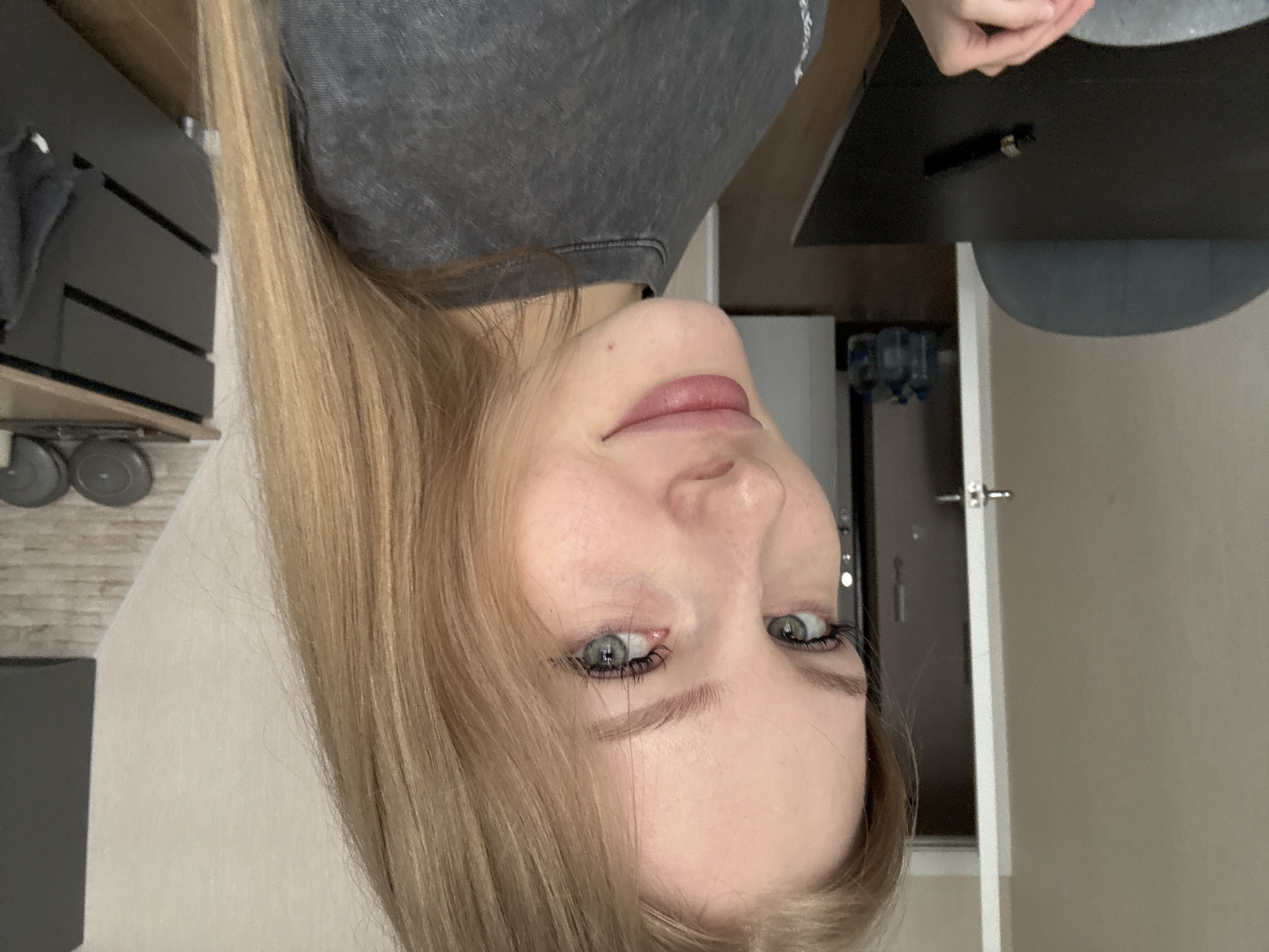 A close-up, rotated, medium indoor shot depicts a fair-skinned, blonde-haired woman with blue eyes. She is wearing a gray t-shirt. Behind her is a desk with a black surface, a gray chair, a partially visible refrigerator, and a light-colored wall with a door. There is also a dark gray shelf with weights on it, and a brick-patterned wall is partly visible. This image was likely taken in a private residence. There is no evidence to support this image was taken in Perm, Russia.