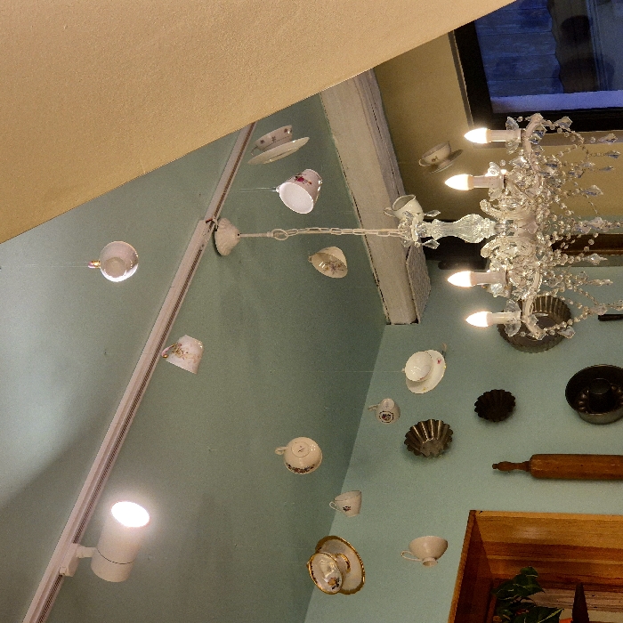The image shows a creative ceiling decoration. Teacups and saucers are suspended on strings, appearing to float in the air. A crystal chandelier provides light and adds elegance. Various baking utensils, including molds and a rolling pin, are mounted on the wall below, enhancing the quirky and whimsical aesthetic. A spotlight shines on the wall, emphasizing the unusual display.