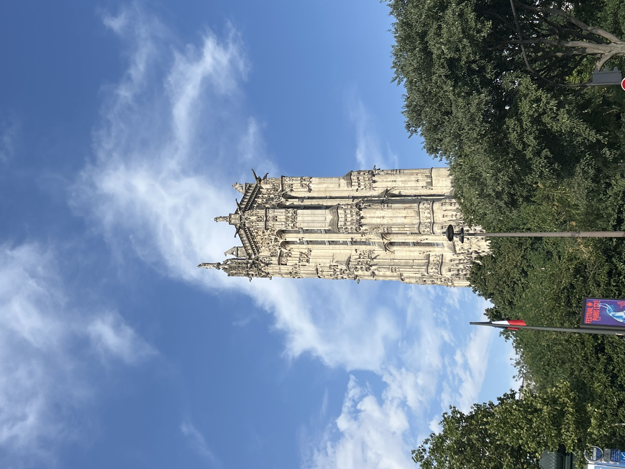 The image shows the Tour Saint-Jacques in Paris, France, under a blue sky with scattered clouds. The tower, a Gothic monument, is centrally located and dominates the upper portion of the frame. It is intricately detailed with carvings and statues, and gargoyles are visible on its upper sections. Lush green trees surround the base of the tower, partially obscuring it from view. In the foreground, a French flag is visible on a flagpole, and a poster with the text "FESTIVAL PARI CITE" can be seen. The scene appears to be a public space on a clear day, with elements suggesting a festival or event taking place.