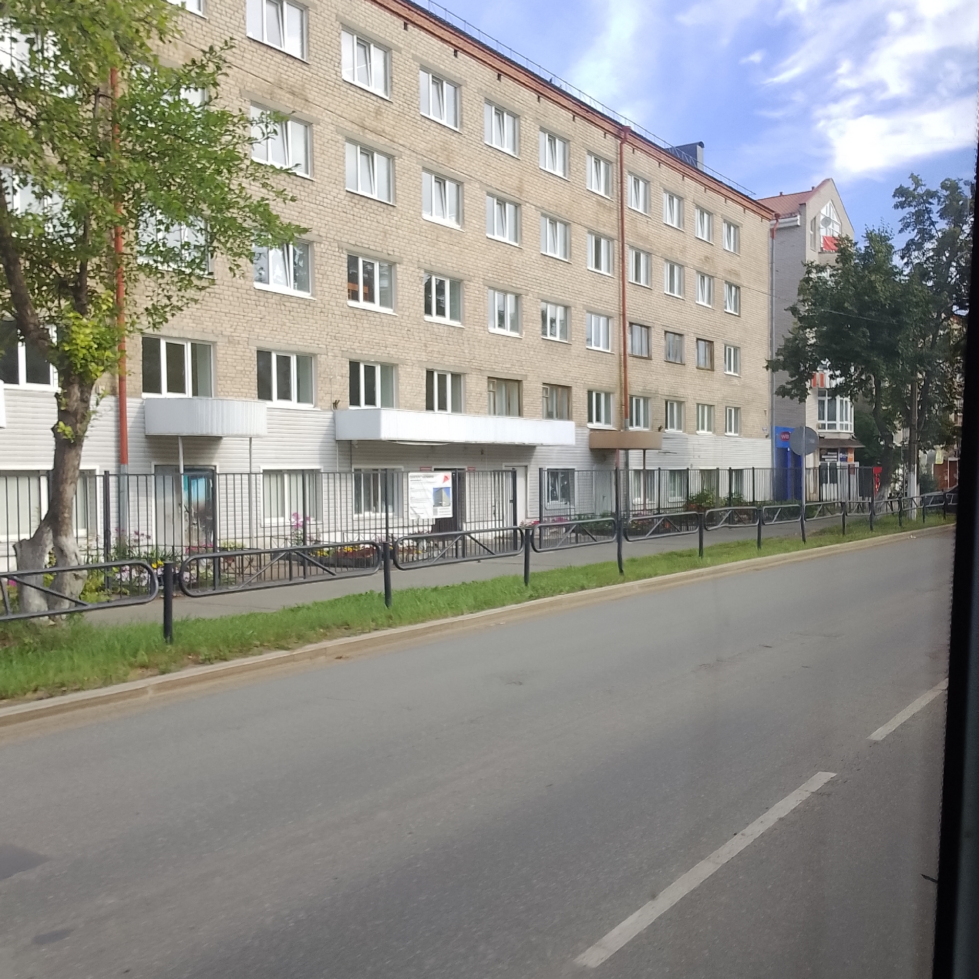 The image shows a long, multi-story brick building on the left side of a paved road. The building has numerous rectangular windows with white frames. A black metal fence runs along the sidewalk in front of the building, separating it from the road. There are some trees and green grass in front of the fence. A small portion of another building with a gabled roof and a window is visible to the right of the main building. The sky above is partly cloudy. The image appears to be taken from a moving vehicle, indicated by the blur of the road and the view through what seems to be a window. The scene is consistent with an urban residential or institutional area. The presence of a sign on the building with Russian text suggests the location is in Russia, and specifically identified as being in the city of Chaikovsky, Russia.