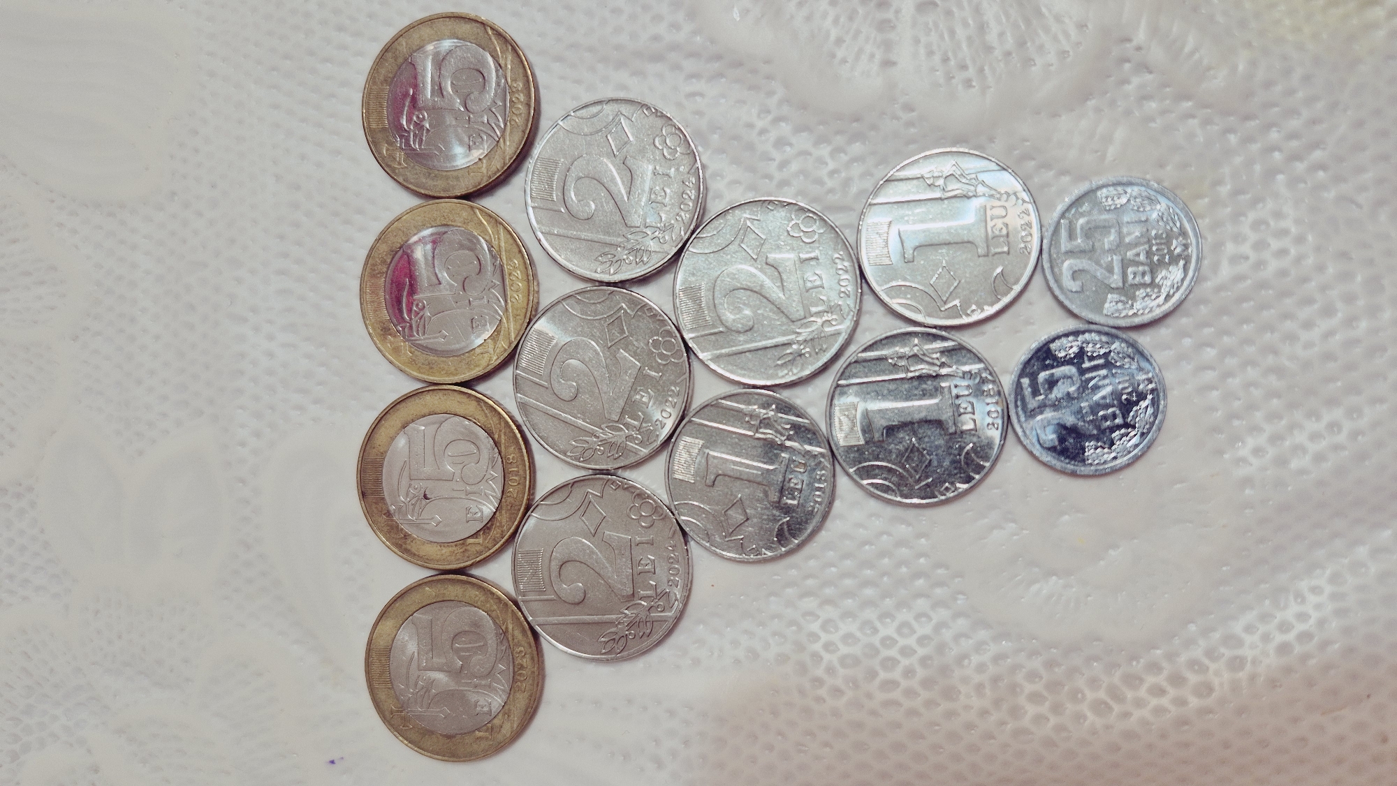 The image shows a collection of Romanian coins arranged in a triangular pattern on a textured, light-colored surface that appears to be a tablecloth. The coins are of various denominations, including 50 bani, 1 leu, and 2 lei. The coins are predominantly silver and gold in color. The lighting in the image is soft, creating a slightly blurred effect. There are no people or animals present in the image. The background is out of focus, but it suggests an indoor setting. The image does not contain any information related to the city of Ialoveni, Moldova.