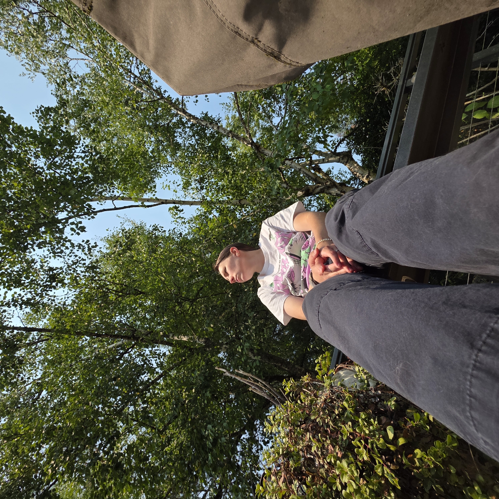 The image depicts a low-angle shot of a person sitting outdoors, looking up at the sky. The person is wearing a white t-shirt with a graphic print and dark pants. Their hands are clasped in their lap, and they are adorned with bracelets. The background consists of lush green trees and a clear blue sky, suggesting a park or natural setting. The presence of a large, textured, brown object on the right side of the frame could indicate a piece of outdoor furniture or equipment. The overall scene is peaceful and relaxed, with the person appearing contemplative. There is no specific information or visual cues that would allow for the determination of the location in Portland, United States of America.