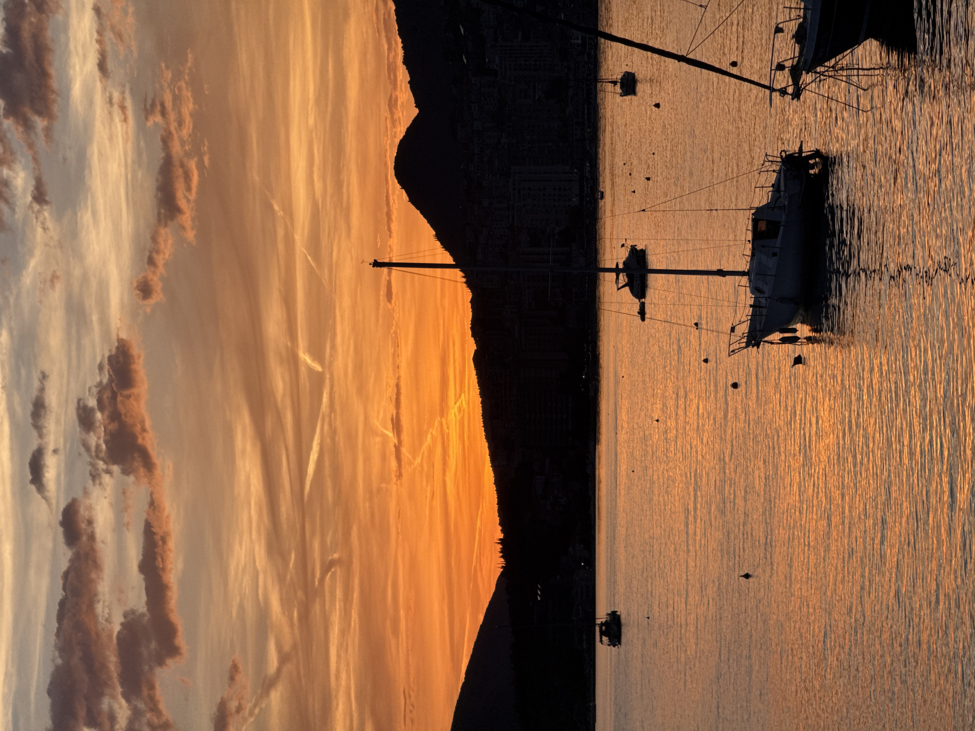 This image depicts a coastal sunset scene over the Bay of Kotor, specifically in the vicinity of Herceg Novi, Montenegro. The sky is dominated by intense orange and yellow hues from the setting sun, with scattered cumulus clouds appearing as dark brown and grey masses in the upper portion. Streaks of light orange and white cirrus clouds are also present, along with several prominent contrails crossing the sky.
A dark silhouette of mountainous terrain spans the entire horizon, rising steeply from the water's edge. Against this silhouetted landscape, a sprawling city skyline, identifiable as Herceg Novi, is visible, its buildings forming dark, angular shapes. Sparse tree lines are discernible on the lower slopes of the mountains.
The water surface reflects the vibrant orange and yellow light of the sunset, creating a shimmering, rippled effect across the bay. Numerous boats are scattered across the water, appearing as dark silhouettes against the glowing reflections. In the mid-ground, slightly left of center, a prominent sailboat with a single mast is anchored, its hull and rigging clearly outlined. Another sailboat is partially visible in the lower right foreground, its mast and rigging dominating that section. Several other smaller boats, including a motorboat on the far left and another sailboat in the mid-right, are present further in the bay. Multiple small, dark buoys are visible floating on the water's surface throughout the scene. There are no discernible people or specific actions visible in the image.