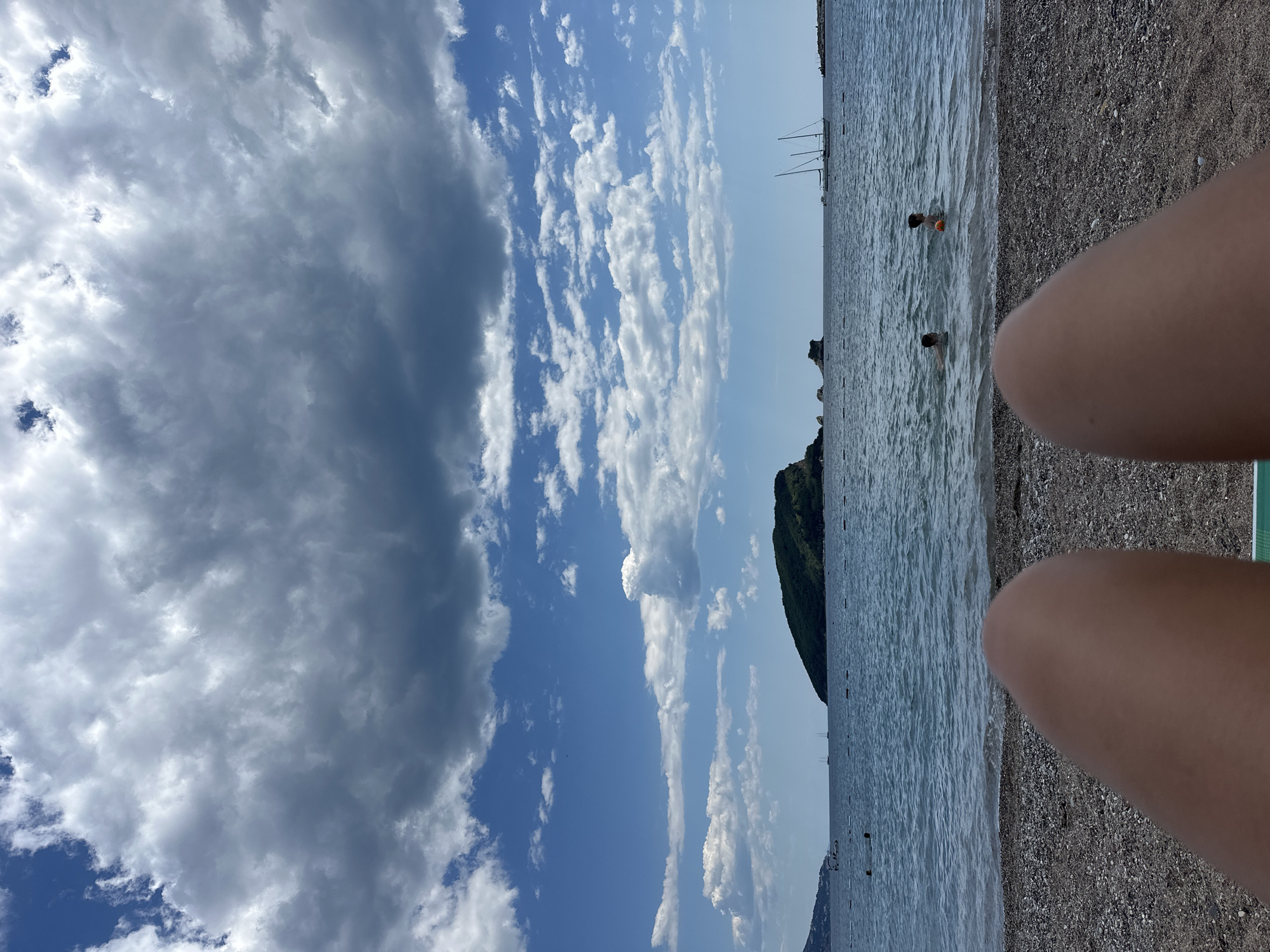 The image depicts a seaside scene in Budva, Montenegro. The view is oriented with the sky and clouds on the left and the Adriatic Sea on the right. In the sea, two people are visible swimming in the water. The beach is pebbly. A green, mountainous landform is visible in the distance. Additionally, two legs of a person are visible in the foreground.