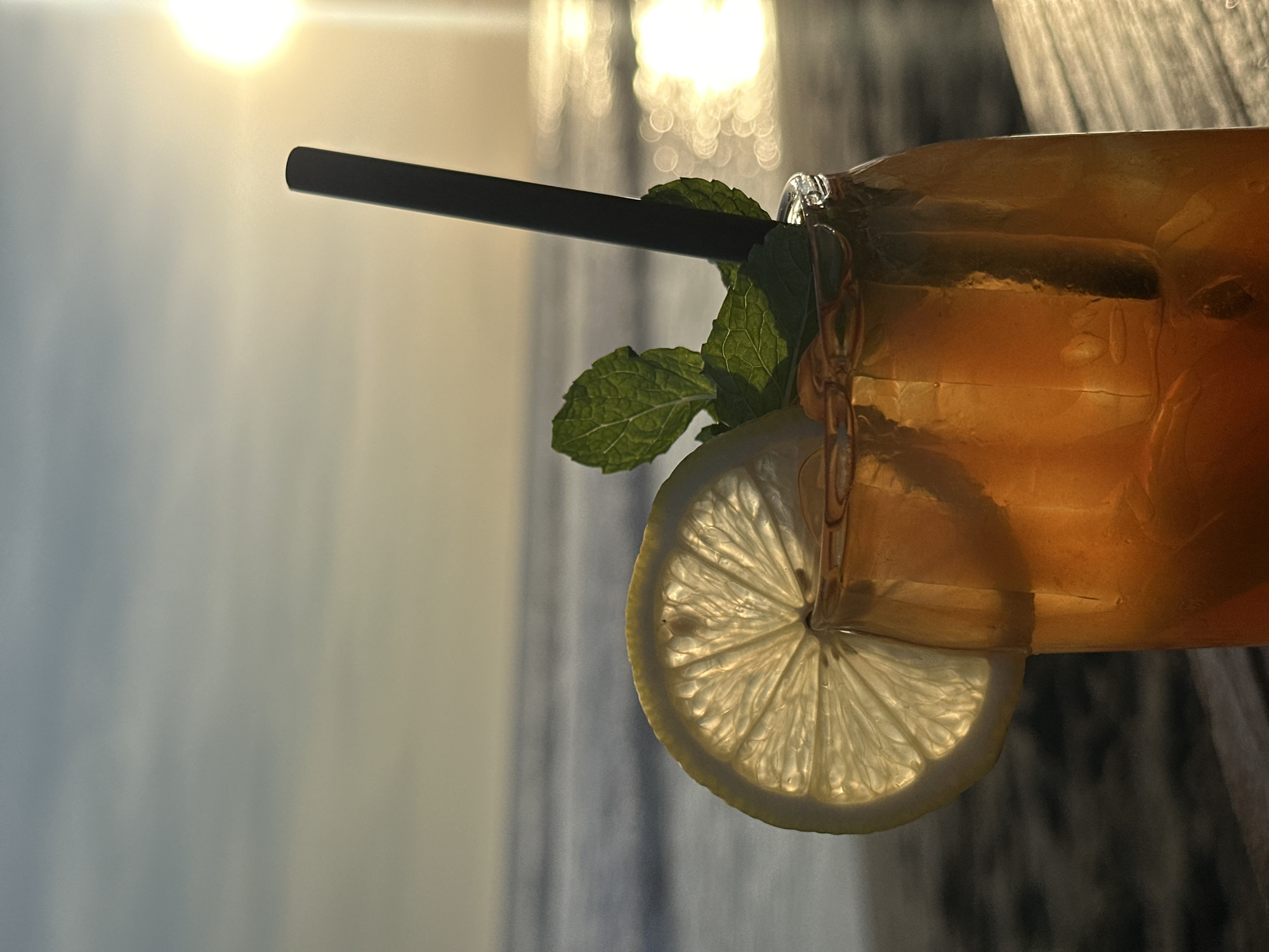 The photograph depicts a glass of iced tea, garnished with a slice of lemon and a sprig of mint, set against a blurred coastal backdrop at sunset. A black straw extends from the glass. The scene is located in North Kuta, Indonesia, suggested by the coastal environment and sunset lighting. The photograph is taken at eye level, with the foreground dominated by the beverage. The lighting is soft and diffused, with the sun low on the horizon. The sea is visible as a blurred expanse in the background.