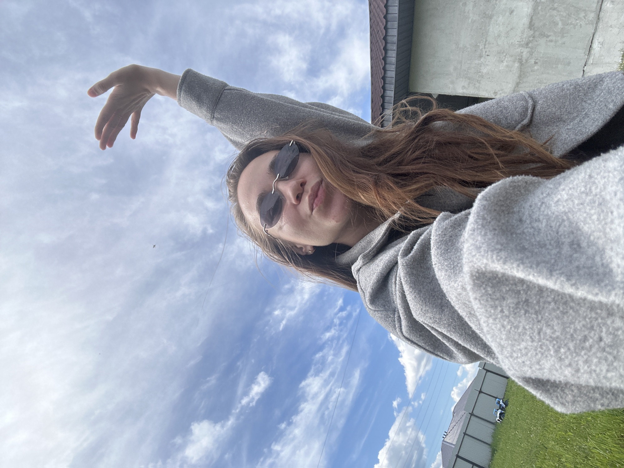 A tilted, low-angle selfie features a young woman outdoors in Kharnavy, Russia. She has long, reddish-brown hair and wears small, dark oval sunglasses. Her face is slightly turned towards the camera, with her lips subtly puckered in a casual expression. She is dressed in a textured grey, long-sleeved top or jacket. Her left arm is extended upwards and to the left, with her hand open and fingers slightly spread, as if posing.
The background is predominantly a bright blue sky filled with scattered white clouds, suggesting daytime with fair weather. In the upper right, part of a building with a brown tiled roof and a light-colored wall is visible. The lower right shows green grass, a tall grey metal fence with vertical panels, and faint power lines stretching across the sky. A small, white and blue object, possibly a toy, rests on the grass near the fence. A tiny, dark shape, likely a bird, is high in the sky on the left. The scene captures a relaxed moment outdoors. No text is visible.