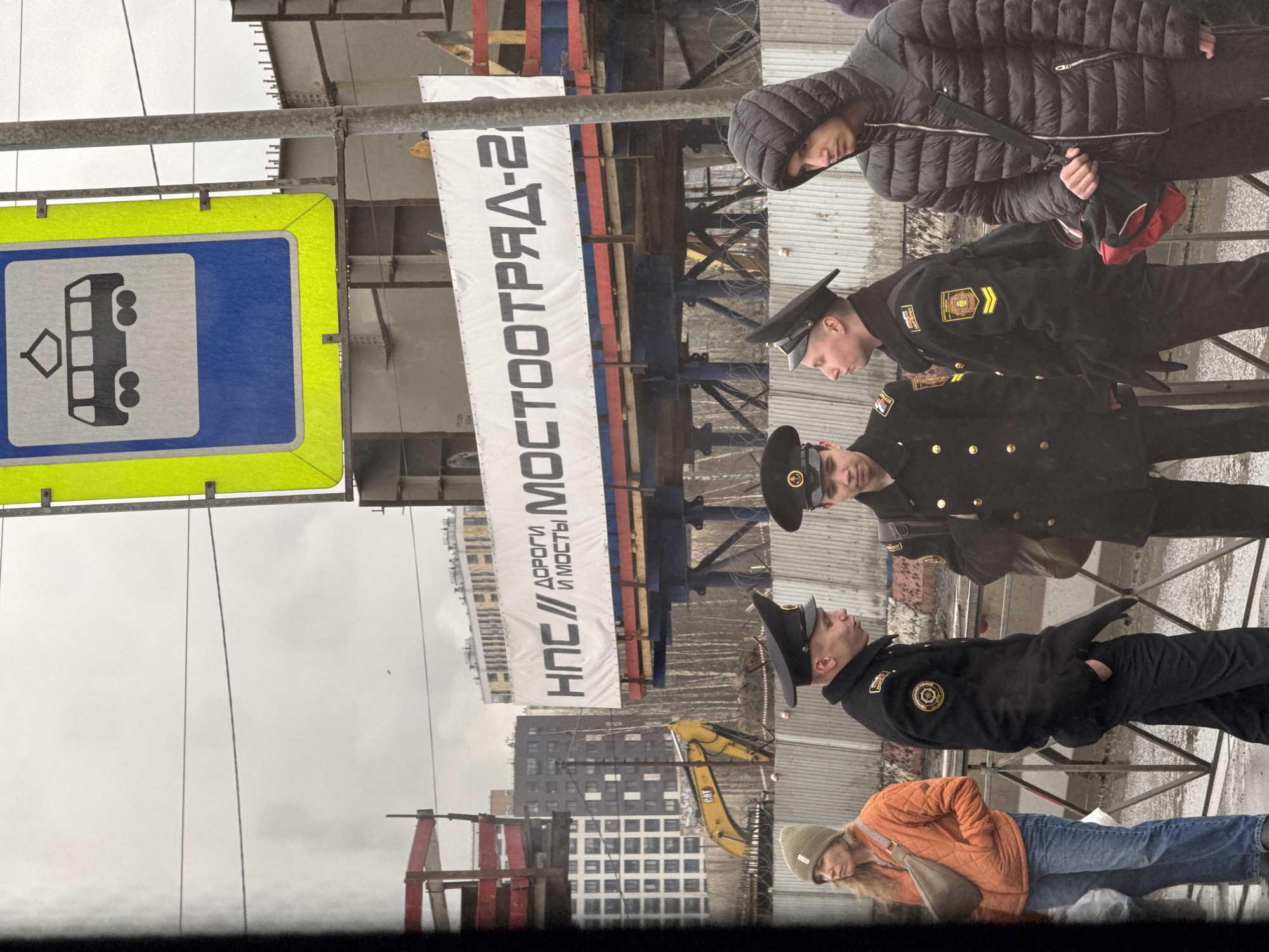 The image shows a street scene in Saint Petersburg, Russia. Three men in dark uniforms with hats stand on a sidewalk. A woman in a puffy black jacket is also present, along with another woman in an orange jacket and jeans. In the background, there is a construction site with scaffolding, an excavator, and a banner written in Russian, НПС// ДОРОГИ и МОСТЫ// МОСТООТРЯД-2. A blue and yellow public transportation sign is visible on a pole. Modern buildings are visible beyond the construction site. The sky is overcast.