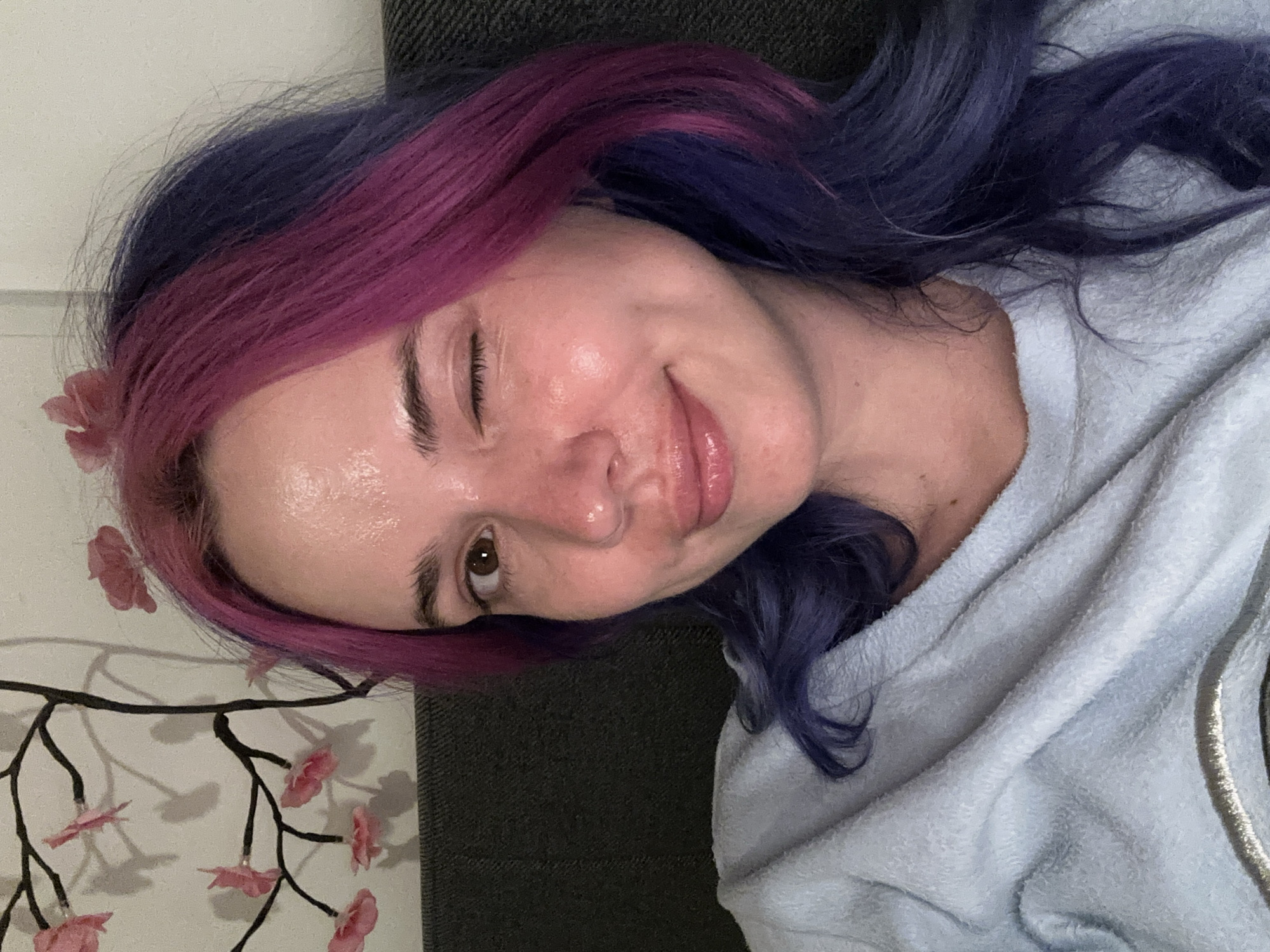 The media file features a headshot of a woman lying down indoors. She has striking two-toned hair, dyed vibrant pink/magenta on the front and darker blue/purple towards the back. The woman is winking with her right eye closed and has a slight, relaxed smile, conveying a playful and friendly emotion. Her skin has a natural, subtle sheen.
She is wearing a light blue, soft-textured garment, which appears to be a fleece hoodie or robe, with a visible zipper detail on the right side. The background behind her head is a dark grey/black textured surface, possibly a pillow or couch. To her left, there is a white wall adorned with a decorative element: a dark, stylized branch featuring several delicate pink flowers, which cast soft shadows on the wall. The lighting is soft and even, characteristic of an indoor environment. There is no visible text in the image.