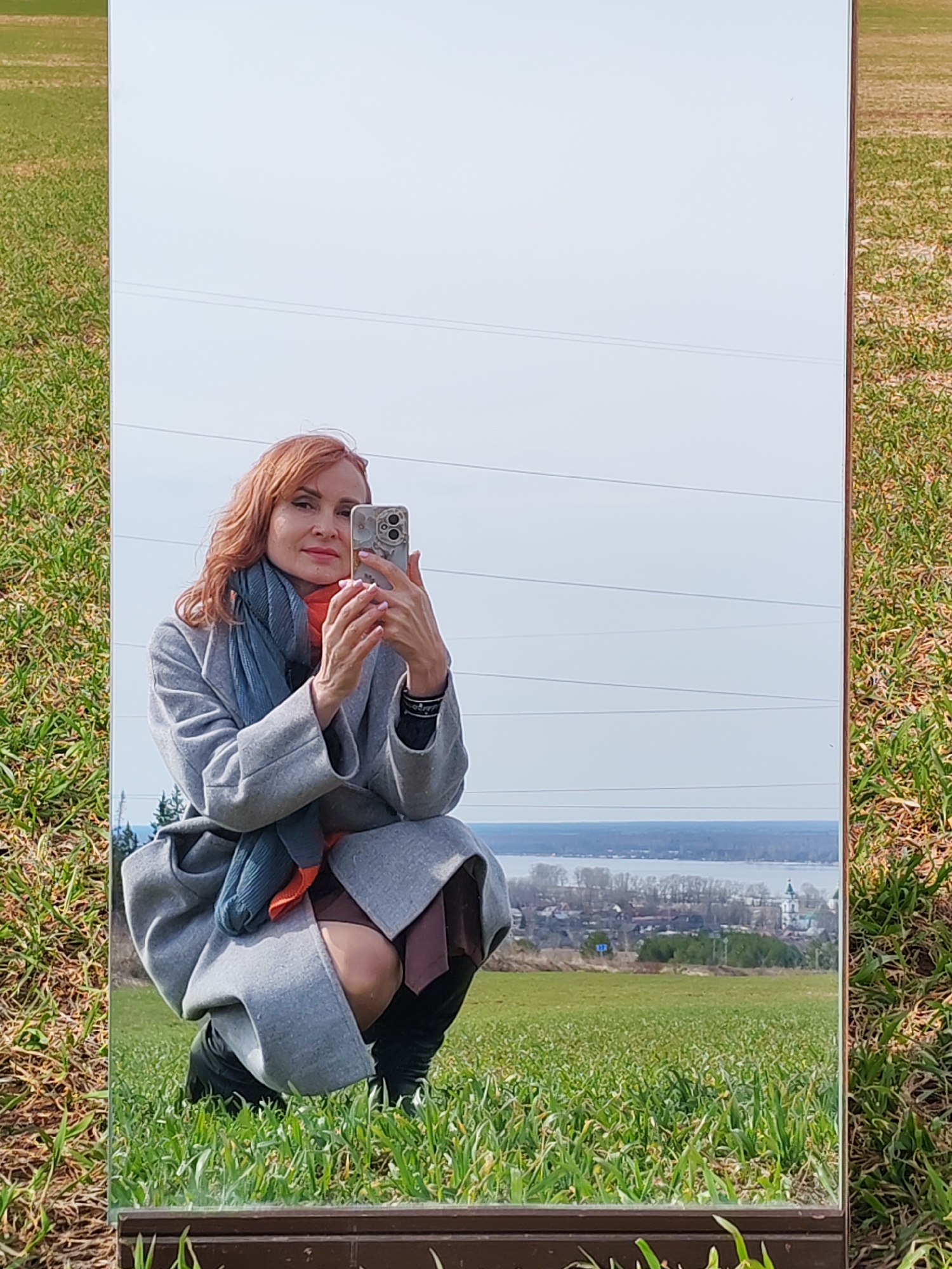 A woman with reddish hair is squatting in a field of green grass, taking a selfie with her phone in a mirror. She is wearing a grey coat, a blue scarf, and a brown skirt. The background of the mirror shows a field, a town, a lake, and a cloudy sky. Power lines are visible in the upper portion of the reflection. The town in the background is Нечкино, Russia.