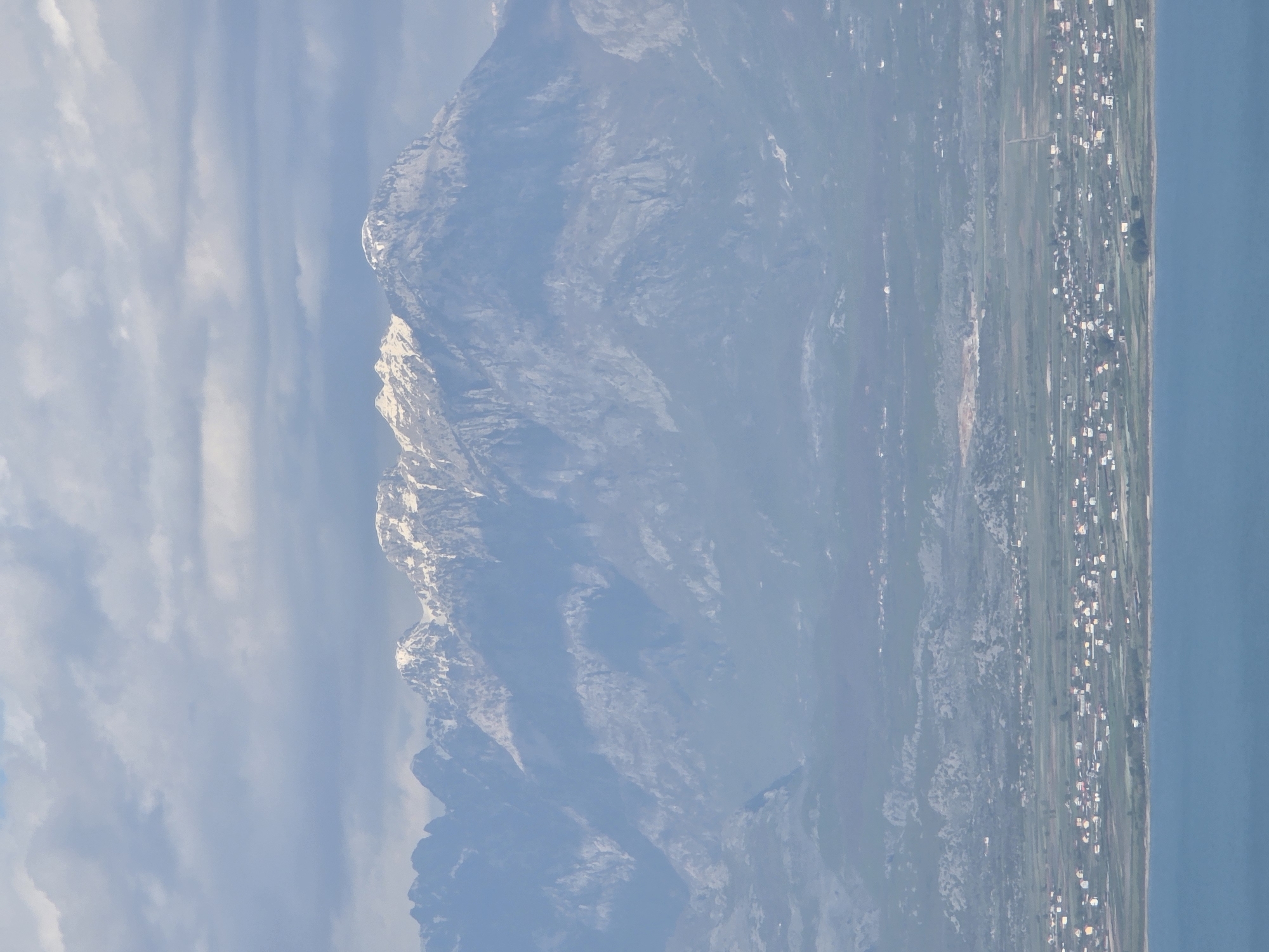 The image shows a landscape with a large mountain range in the background. The peaks are partially covered with snow. In the foreground, there is a coastal plain with vegetation and a settlement consisting of many buildings. The scene may be located near the city of Arbnež, Montenegro, as per the prompt's location constraint. To the right, the plain meets a body of water, likely the Adriatic Sea. The sky is overcast with clouds, contributing to a diffused and somewhat hazy lighting.