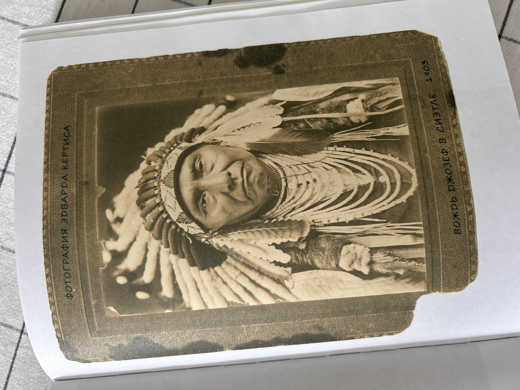 This is a sepia-toned photograph of a Native American man adorned in traditional regalia, including a feathered headdress and beaded necklaces. The man is looking directly at the camera with a neutral expression. The photograph is mounted on a cardboard backing that has decorative borders.
Visible text on the left side of the photograph reads, "ФОТОГРАФИЯ ЭДВАРДА КЕРТИСА". On the right side, the text reads, "ВОЖДЬ ДЖОЗЕФ В СИЭТЛЕ, 1903". This indicates that the photograph was taken by Edward S. Curtis and depicts Chief Joseph in Seattle in 1903. The image appears to be part of a larger collection, possibly an album, given its placement within what looks like a book. The condition of the photograph shows signs of age, with some spotting and wear on the edges of the mount.