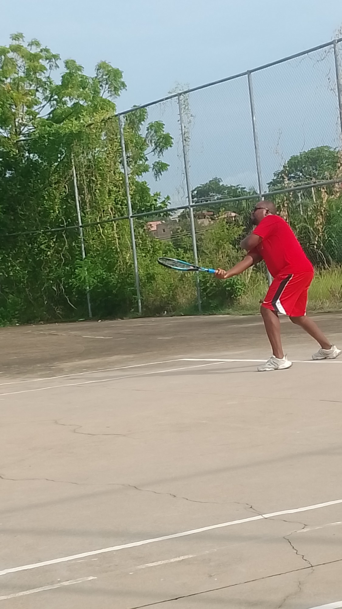 The image depicts an adult male playing tennis on an outdoor hard court in Lokoja, Nigeria, under a bright daytime sky. The man, wearing a red short-sleeved t-shirt, red shorts with black and white accents, white sneakers, and dark-framed glasses, is captured mid-swing in what appears to be a forehand stroke. He is holding a blue and black tennis racket, intently focused on the game.
The tennis court surface is a light grey or beige, showing visible cracks and white lines delineating court boundaries. Behind the court, a tall chain-link fence provides a backdrop. Beyond the fence, there is a dense growth of lush green trees and bushes, indicating a natural or semi-wild environment. In the far distance, through the foliage and fence, some residential or other buildings are faintly visible.
The lighting suggests it is daytime, with bright, clear conditions, and soft shadows are cast on the court. The man's posture conveys concentration and engagement in his athletic activity. No visible text is present in the image. The scene portrays a moment of recreation or practice on a well-used local tennis court.