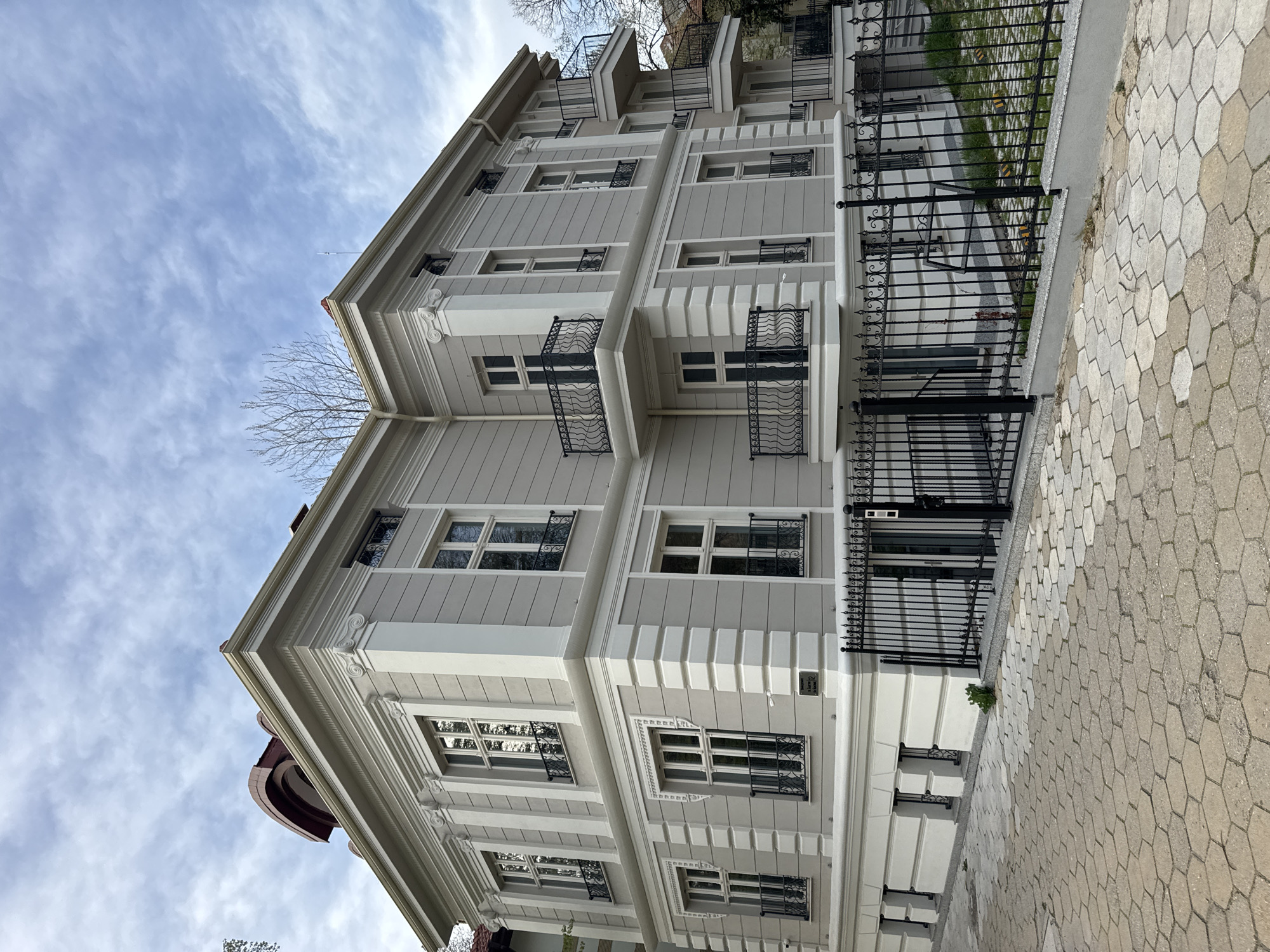 The image depicts the exterior of a building in Plovdiv, Bulgaria, characterized by a light gray facade with white trim and decorative elements. The building has multiple floors with rows of windows, most adorned with wrought iron grilles, and small balconies with ornate railings. A black iron fence runs along the front of the property, separating it from a paved sidewalk composed of hexagonal stones. The sky is visible in the background with scattered clouds. There are no people visible.
