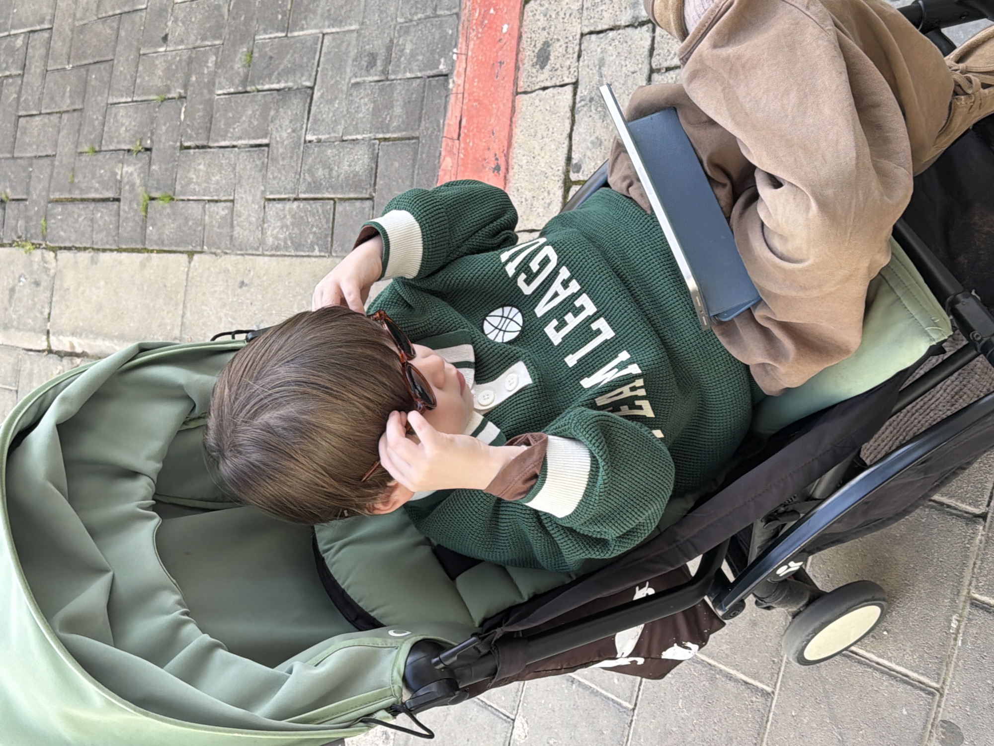 A toddler sits in a light green stroller on a Tel-Aviv sidewalk with cobblestone pavement. The stroller is positioned on the right side of the image, with a red curb running along the top. The child wears a dark green sweater with white lettering. A brown jacket is draped over the toddler’s legs, along with a blue tablet-like object. The child is holding sunglasses near their face.
