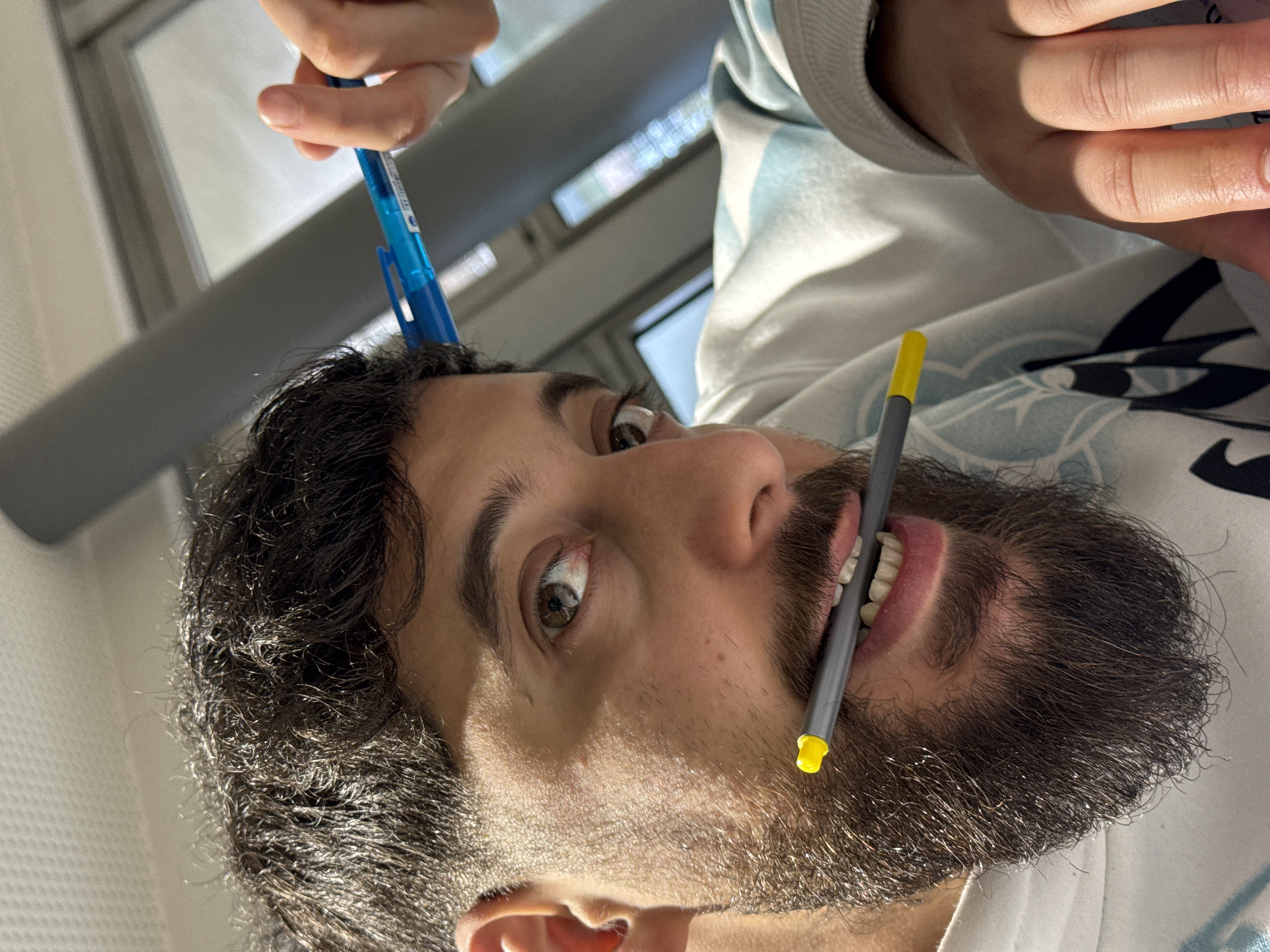 This image, captured indoors in Strasbourg, France, presents a close-up view of a male individual with a secondary hand visible. The primary subject has dark, curly hair, a full dark beard, and brown eyes, directed towards the viewer. He holds a grey pen with a yellow end horizontally between his upper and lower teeth. He is dressed in a light-toned sweatshirt featuring a light blue, patterned design. In the upper right of the frame, a second person's hand holds a blue retractable pen, positioned above and slightly behind the primary subject's head. The background consists of a light-colored ceiling, a prominent grey beam, and a blurred window or reflective surface revealing ambient light. The scene is illuminated by strong, directional light, casting defined shadows on the primary subject's face.