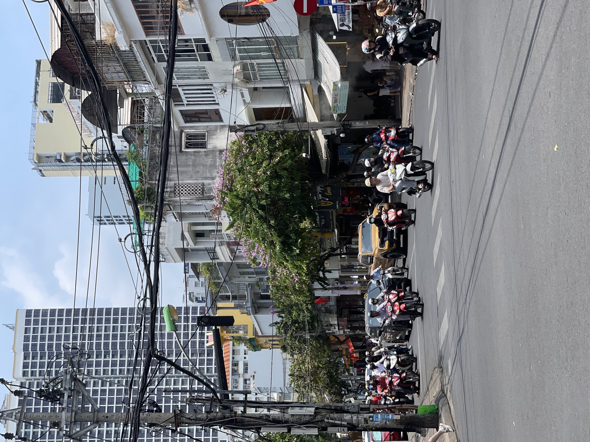 This street-level photograph shows a busy street in Nha Trang, Vietnam. Many motorbikes, each with a rider, are stopped at a crosswalk. The building on the left is a high-rise, and the rest are three- to five-story buildings. Tangled electrical wires are highly visible in the foreground. The sky is partly cloudy, suggesting it is daytime. The crosswalk suggests that vehicles may be about to start crossing in the next few seconds. There is a No Entry sign on the top right.
